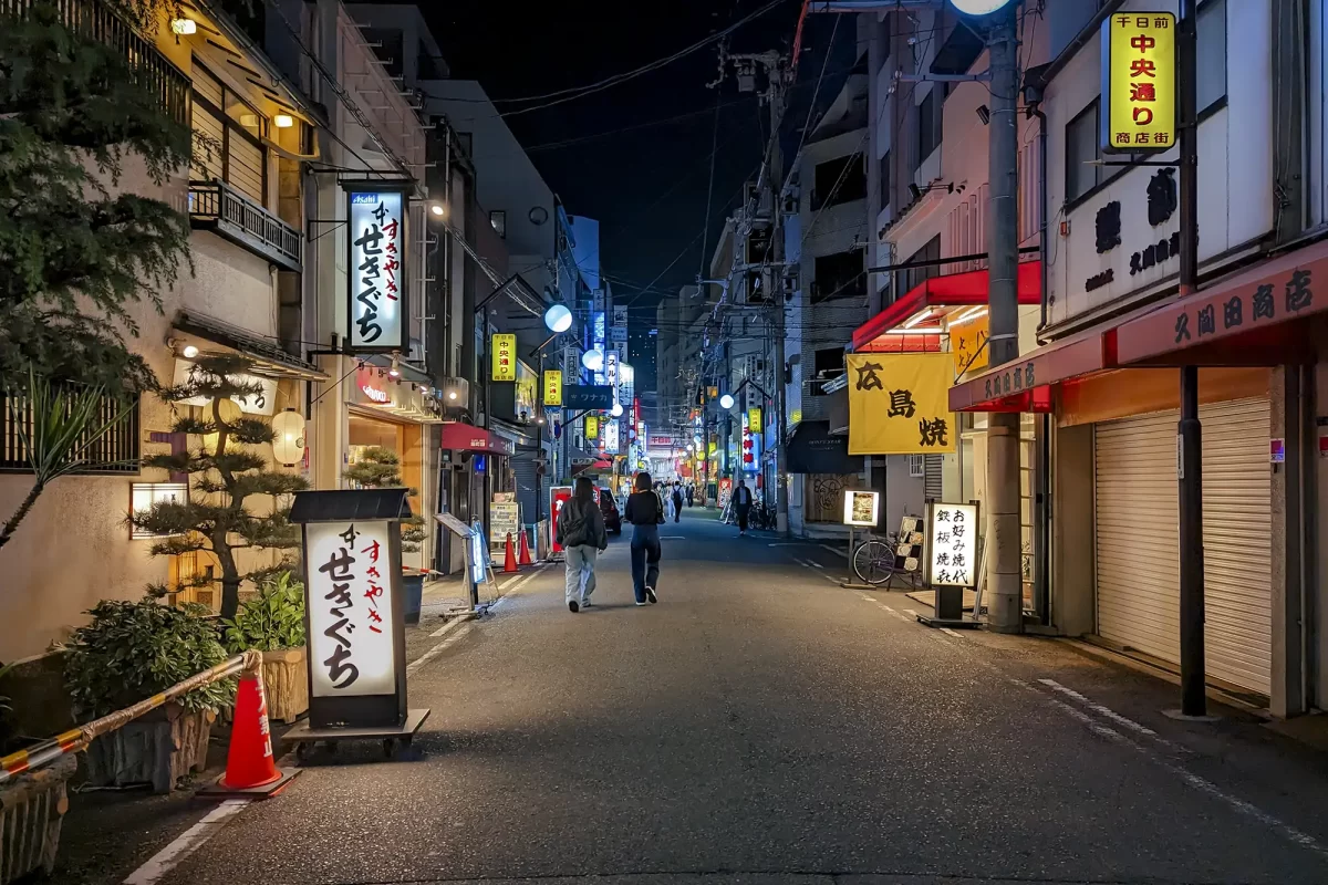 A picture of a narrow street with neon sights and people walking down the street in the city of Osaka, Japan, at night