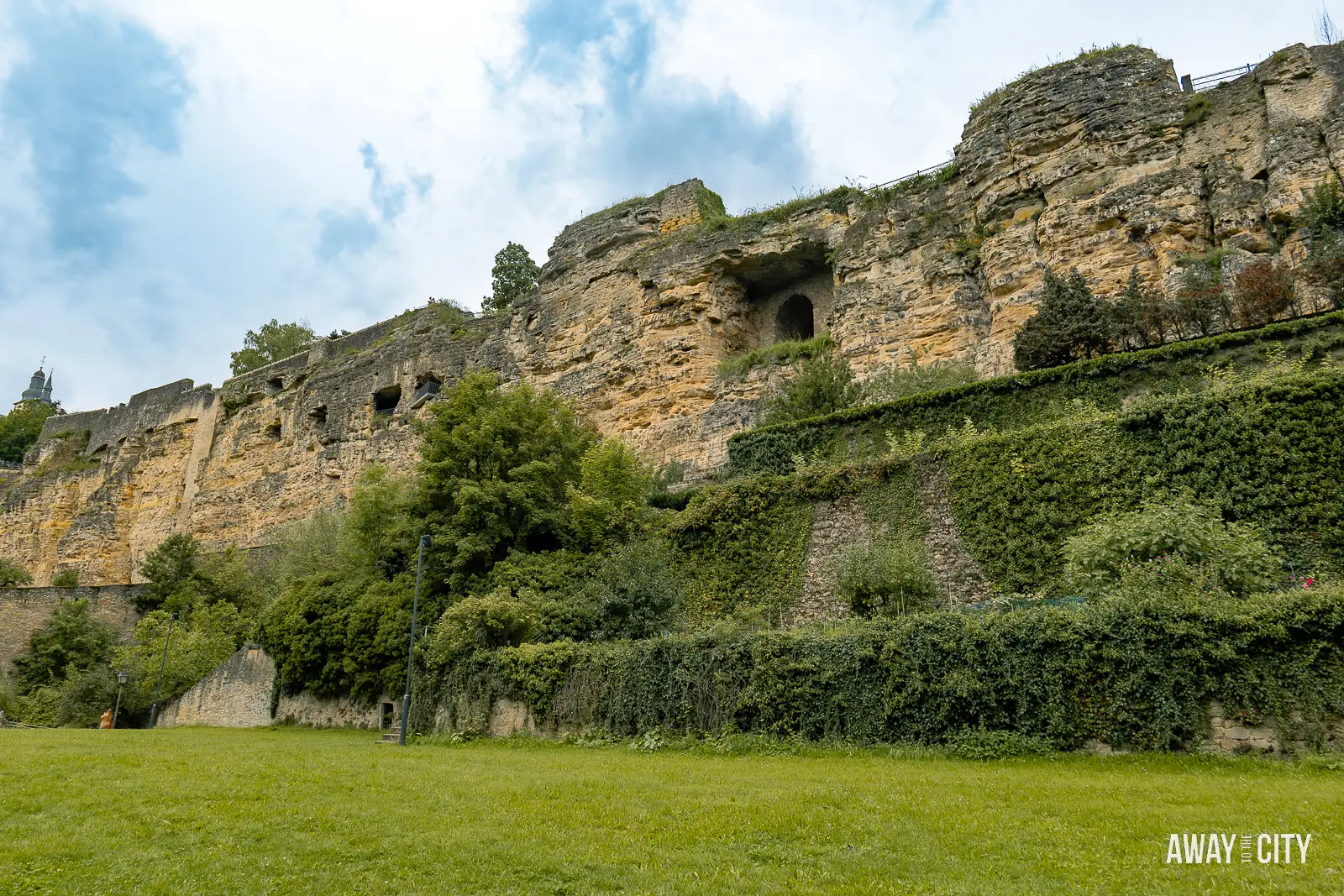 A scenic view of the old quarters and fortifications of the Bock Casemates in Luxembourg City, with a lush green field, rock wall, and trees