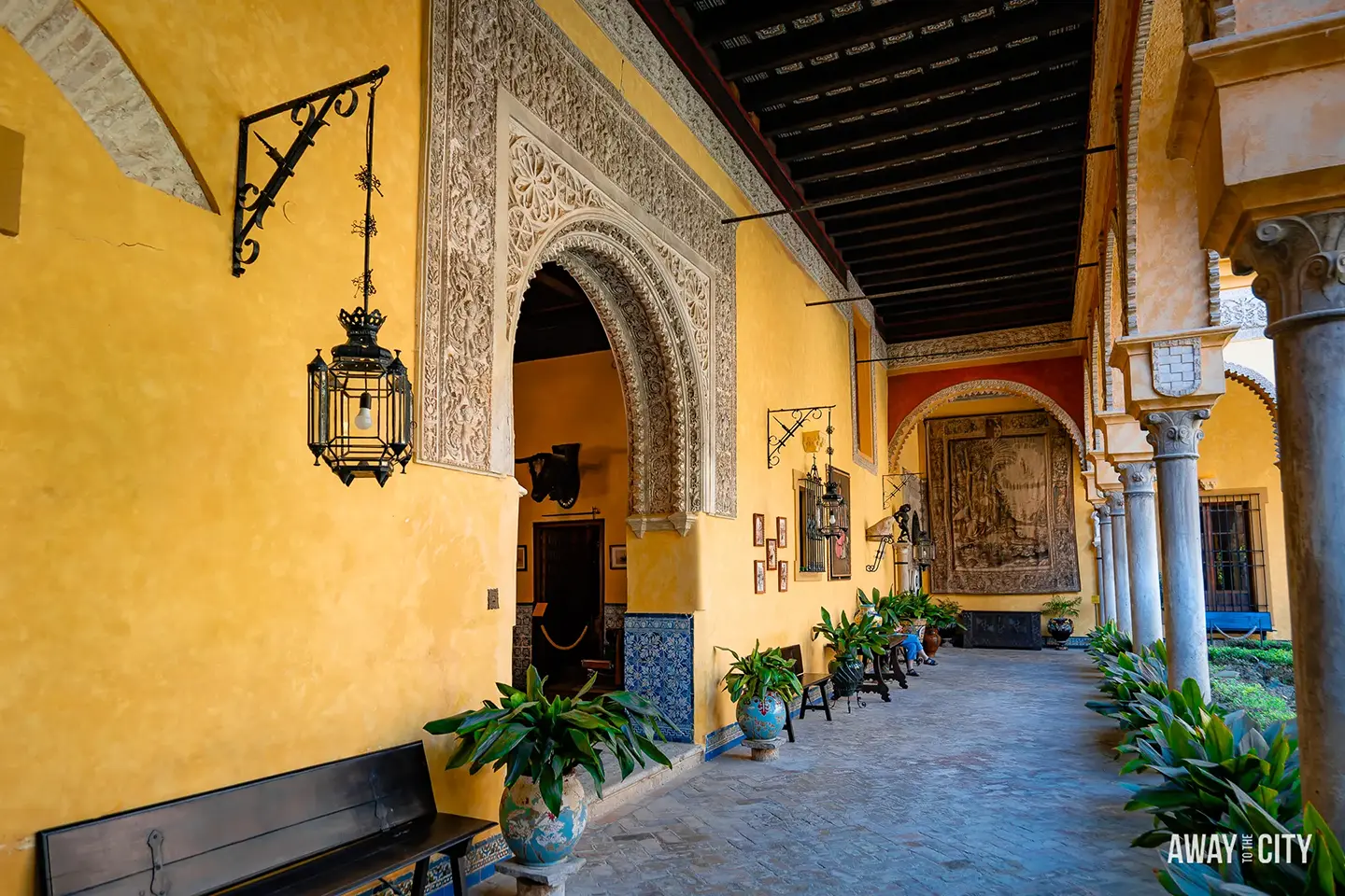 A picture of the yellow, covered hallway in the main garden's courtyard of Palacio de las Dueñas in Seville with plants in pots.
