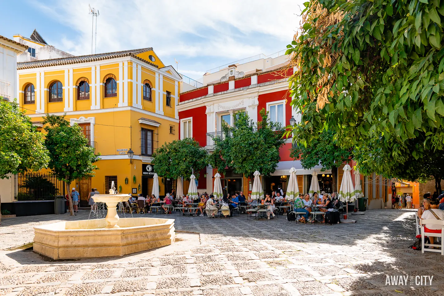 A picture of a small square in the Santa Cruz district of Seville, featuring a fountain and people enjoying lunch at outdoor restaurants.