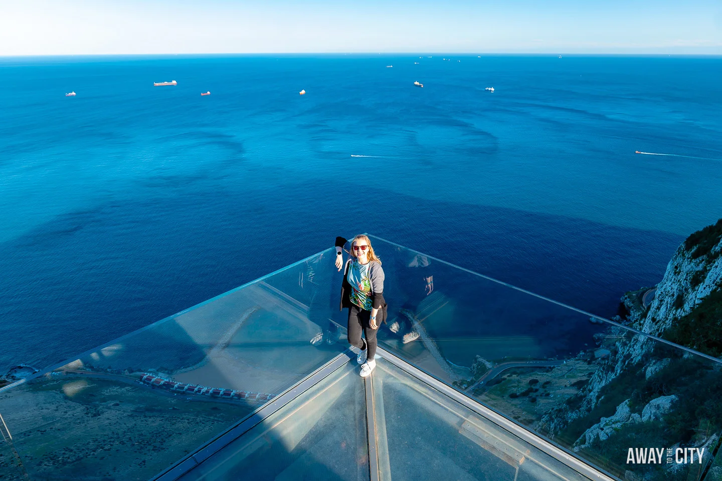 A picture of a person standing on a glass surface of The Skywalk at Gibraltar Rock overlooking the ocean, providing a thrilling and transparent perspective of the sheer drop below.
