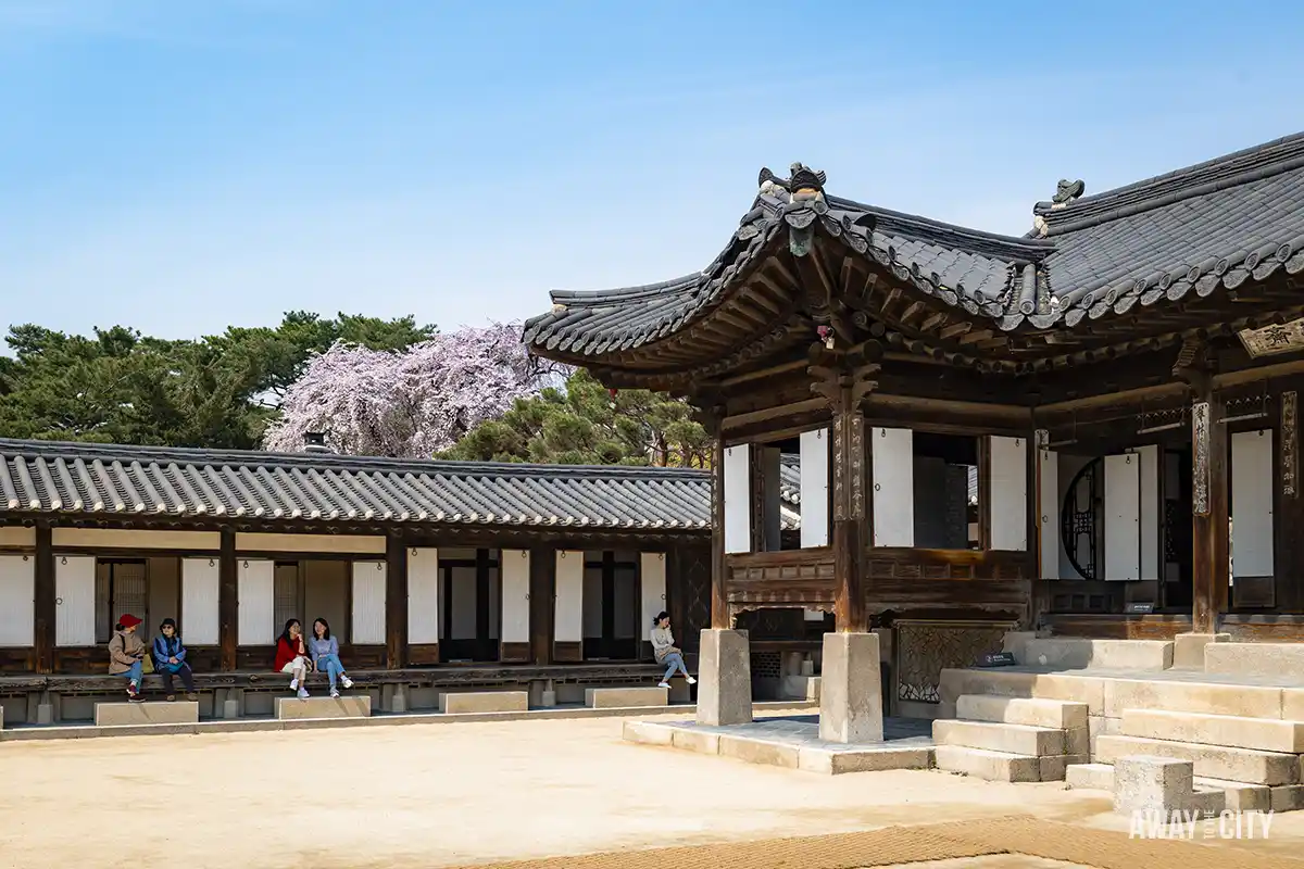 A brown wooden building with a roof and people sitting in front of the building, taken at the Nakseonjae complex of Changdeokgung Palace in Seoul.