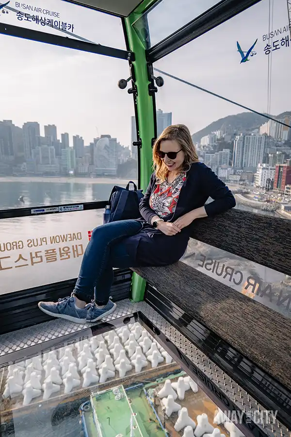 A person sitting on a bench in a cable car with a transparent floor, taken during a Busan Air Cruise ride.