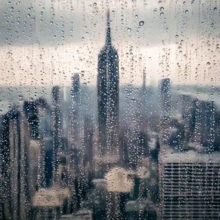 The skyline of New York City on a rainy day viewed through a window with raindrops running down the glass from the heavy pour down.