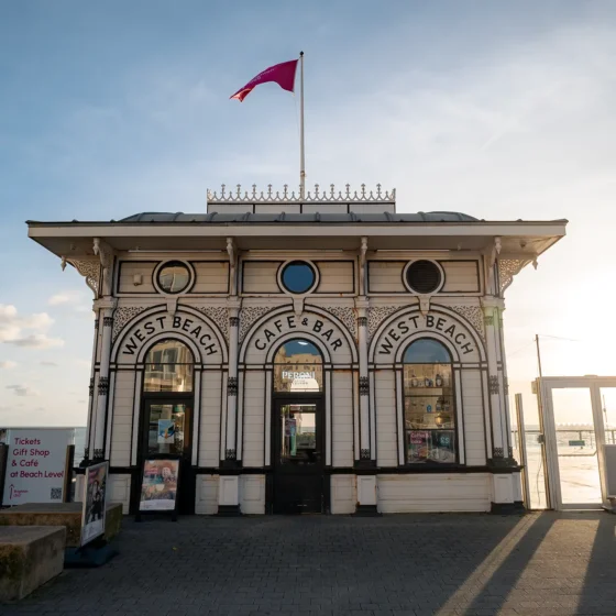 The classic West Beach Café & Bar building on the Brighton Pier, with its distinctive wooden façade and red flag, and the text overlay "Away To The City."