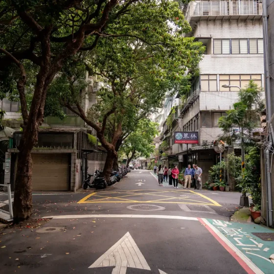 A narrow urban street, shaded by large trees, with pedestrians on the sidewalk, motorcycles parked along the side, and multi-story buildings with balconies and signs, including one with Chinese characters.