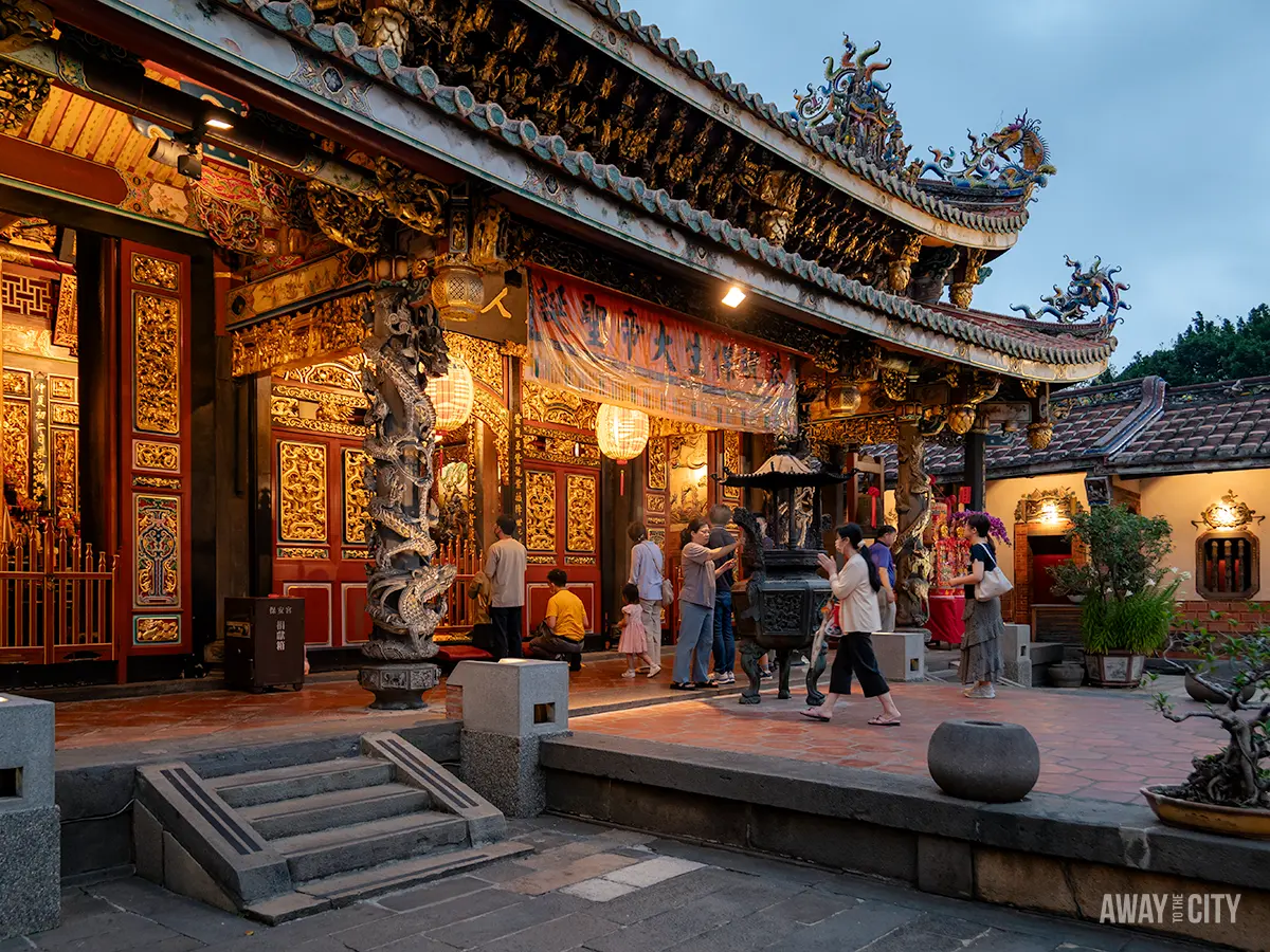 A group of people are visiting a beautifully ornate temple with intricate carvings and decorations, featuring a dragon pillar and traditional lanterns, with the text "Away To The City" in the bottom right corner.