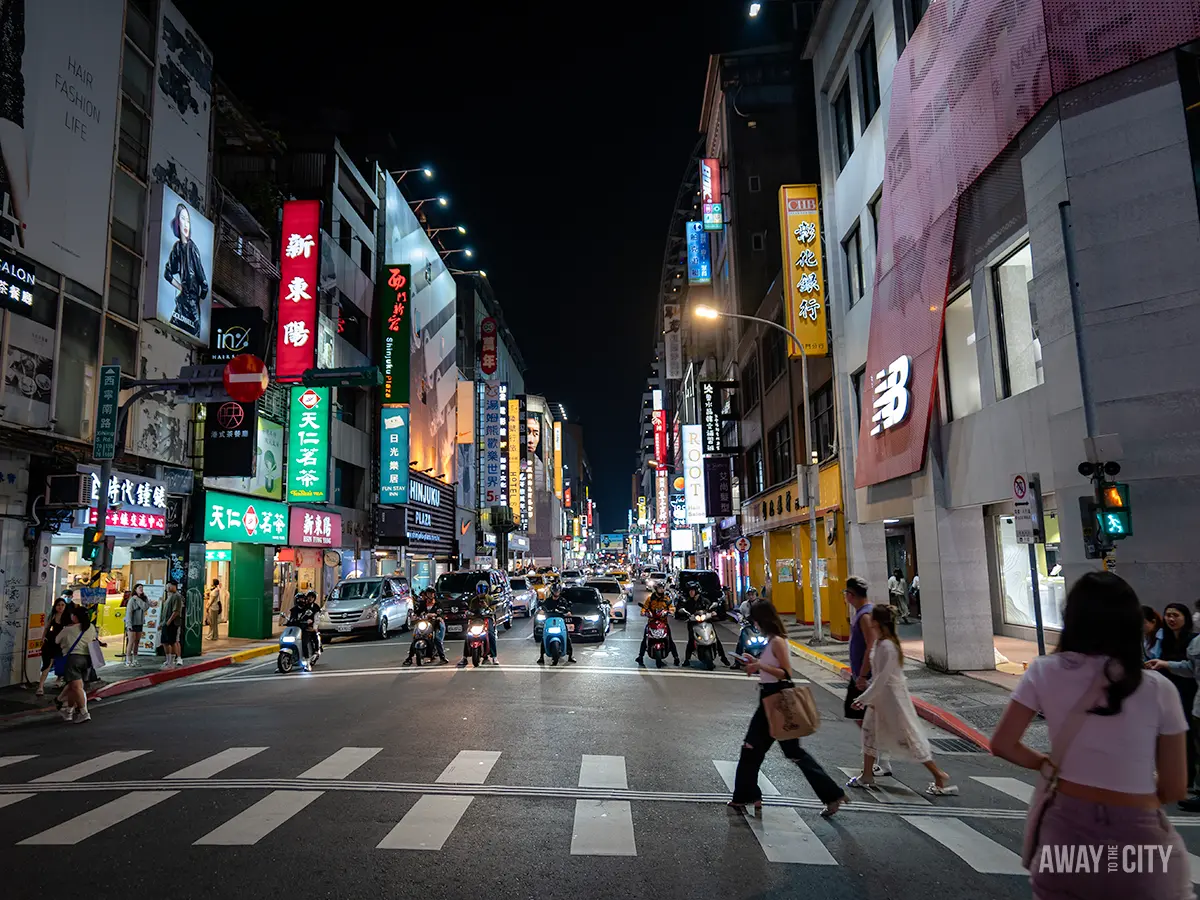 A bustling urban street scene at night, likely in an East Asian city, with tall buildings, bright advertisements, numerous signs with Chinese characters, pedestrians at a crosswalk, scooters and cars at a traffic light, with the text "Away To The City" in the bottom right corner.