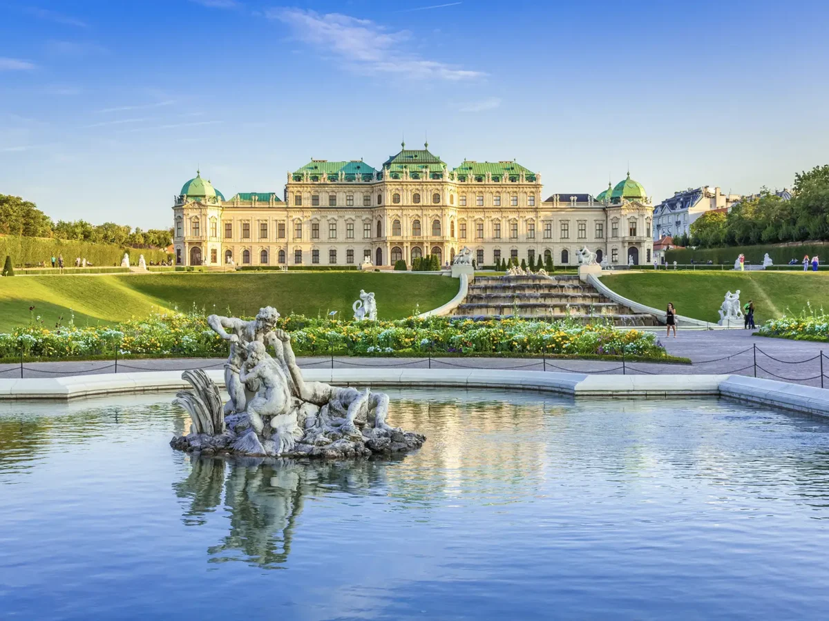An expansive view of Schloss Belvedere in Vienna, a grand palace with a green roof, a terraced garden, and a large reflecting pool with a fountain sculpture in the foreground.