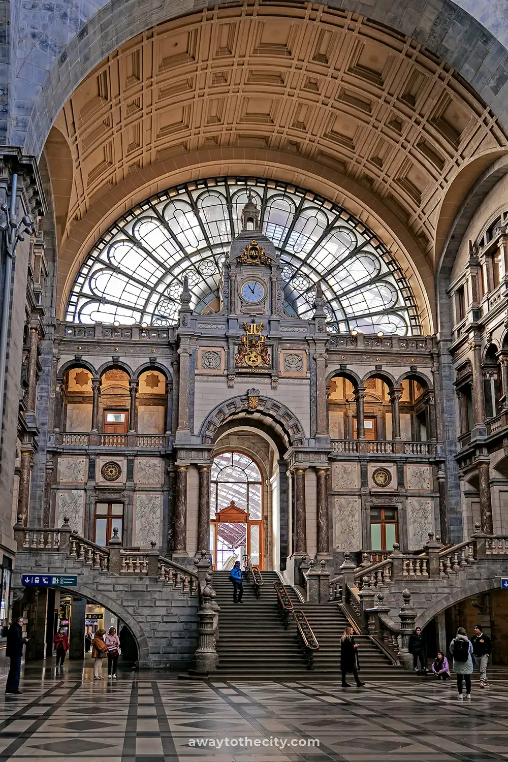 The grand interior of Antwerp Central Station features a large arched window, an ornate clock, and sweeping stone staircases.