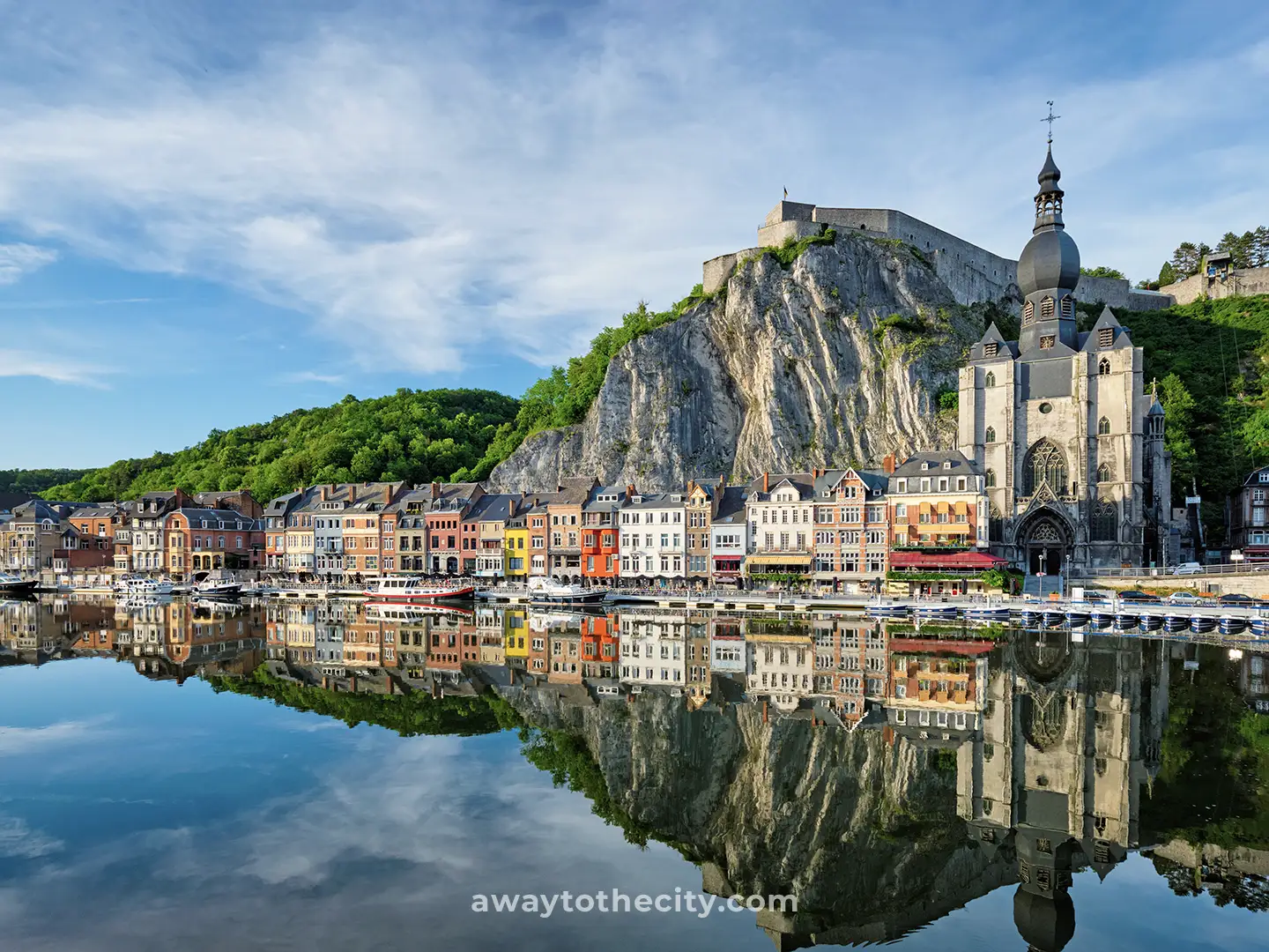 The picturesque city of Dinant, Belgium, with its colourful houses and a large church reflected in the calm river, sits beneath a towering cliff and citadel.
