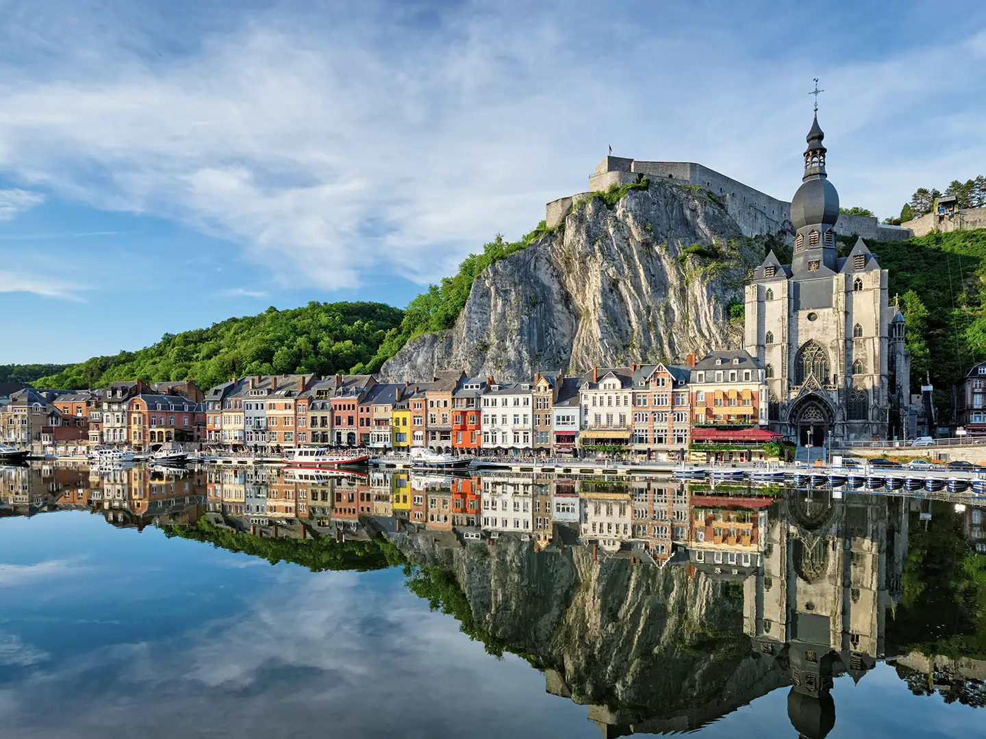 The picturesque city of Dinant, Belgium, with its colourful houses and a large church reflected in the calm river, sits beneath a towering cliff and citadel.