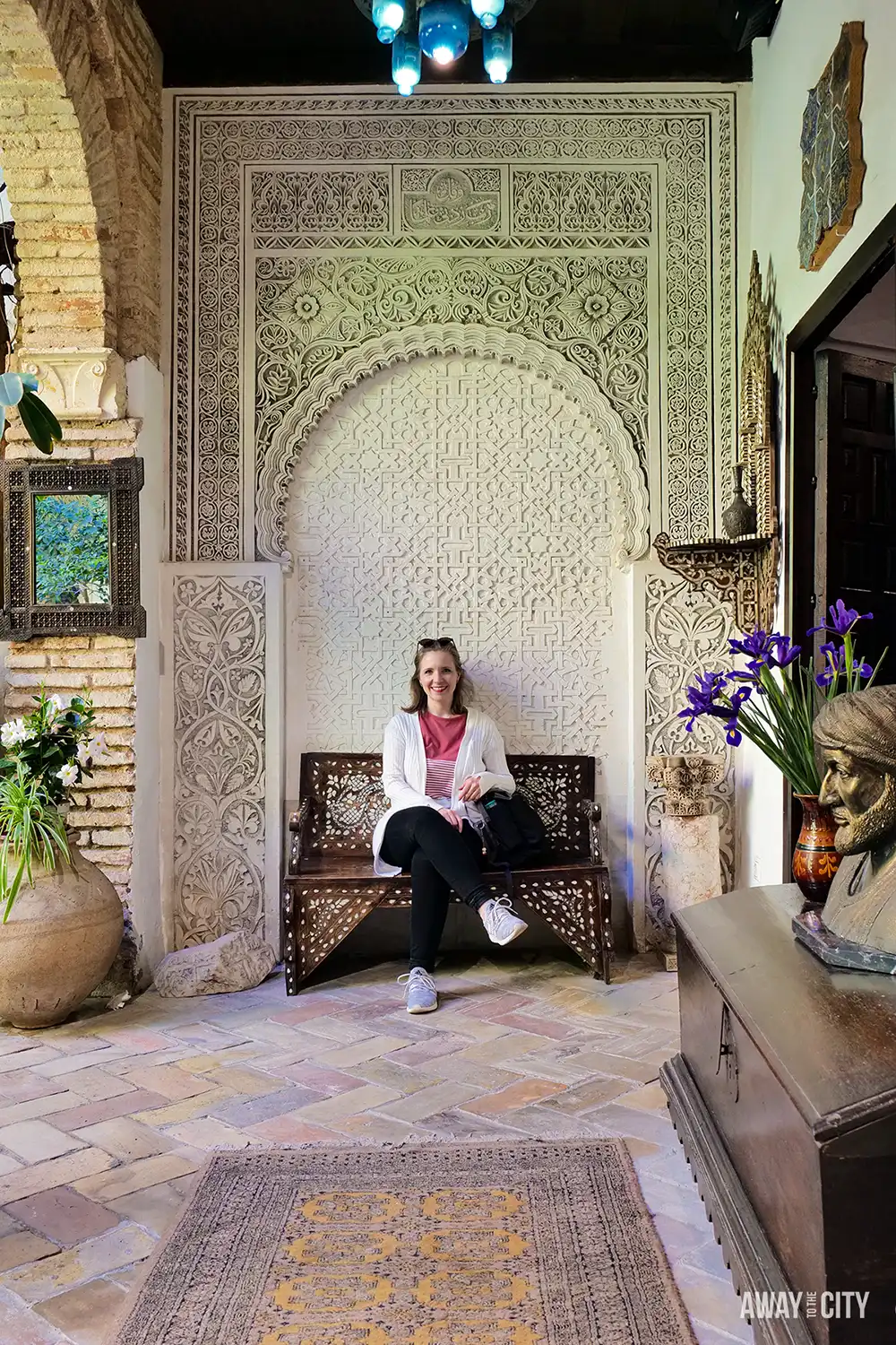 Interior of Casa Andalusi in Cordoba, featuring ornate Moorish arch, intricate carved plasterwork, tiled floor, and traditional decor with plants and antique furnishings.