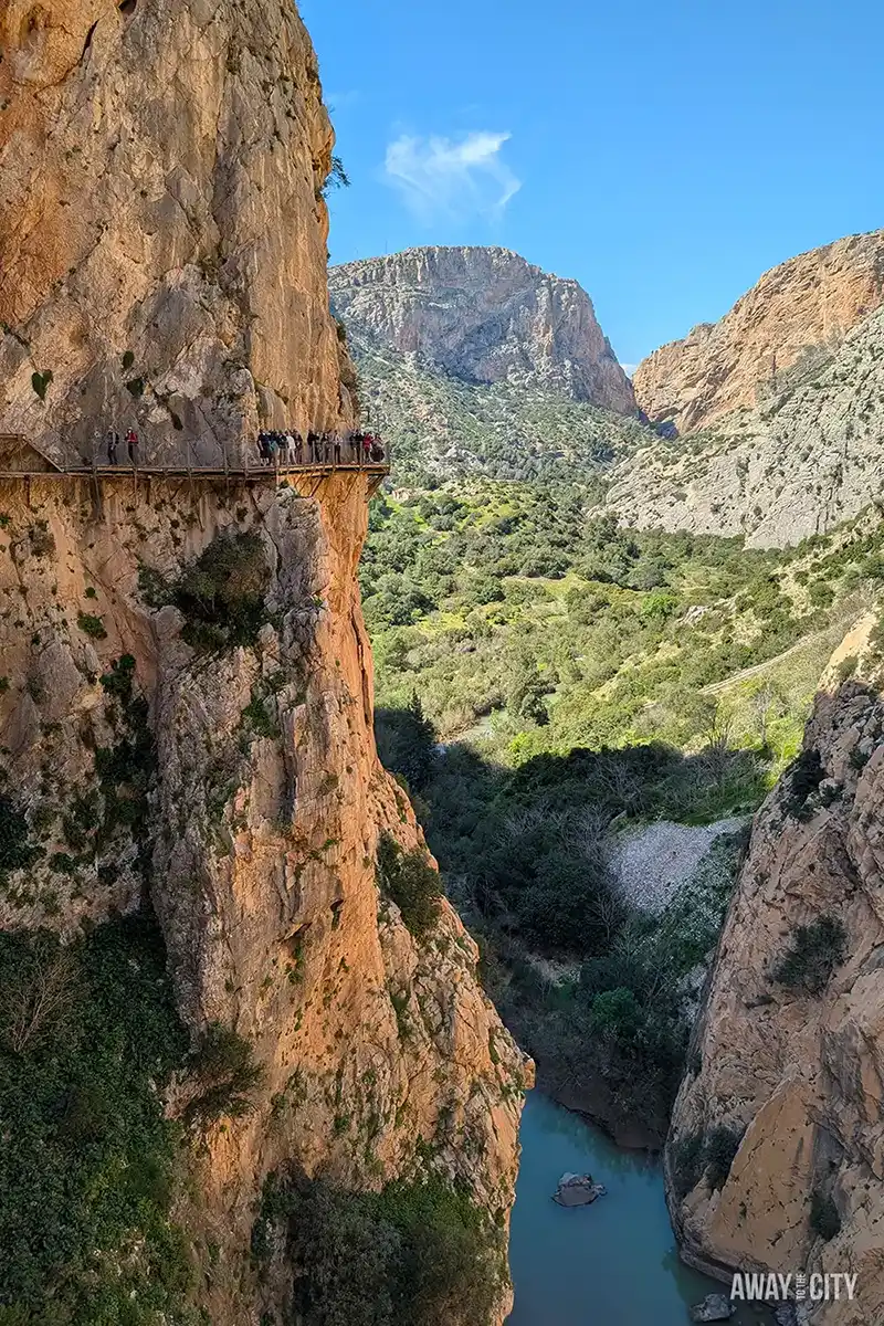 Caminito del Rey walkway clinging to steep gorge cliffs, with visitors crossing a narrow path above a river between rugged mountains under clear blue sky.