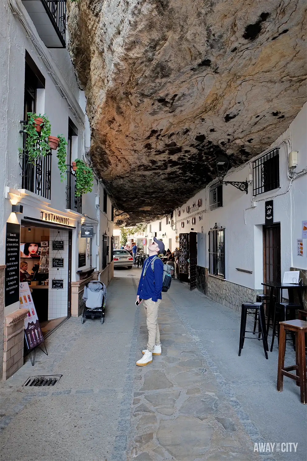 Street in Setenil de las Bodegas with whitewashed houses built beneath an overhanging rock, small shops, and a person standing along the narrow lane.