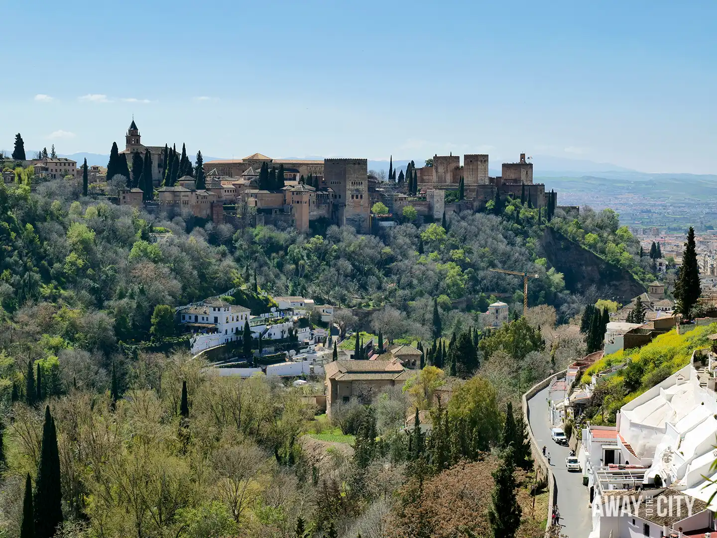 Panoramic view of the Alhambra palace and fortress complex in Granada, perched on a wooded ridge above white houses and winding hillside roads.