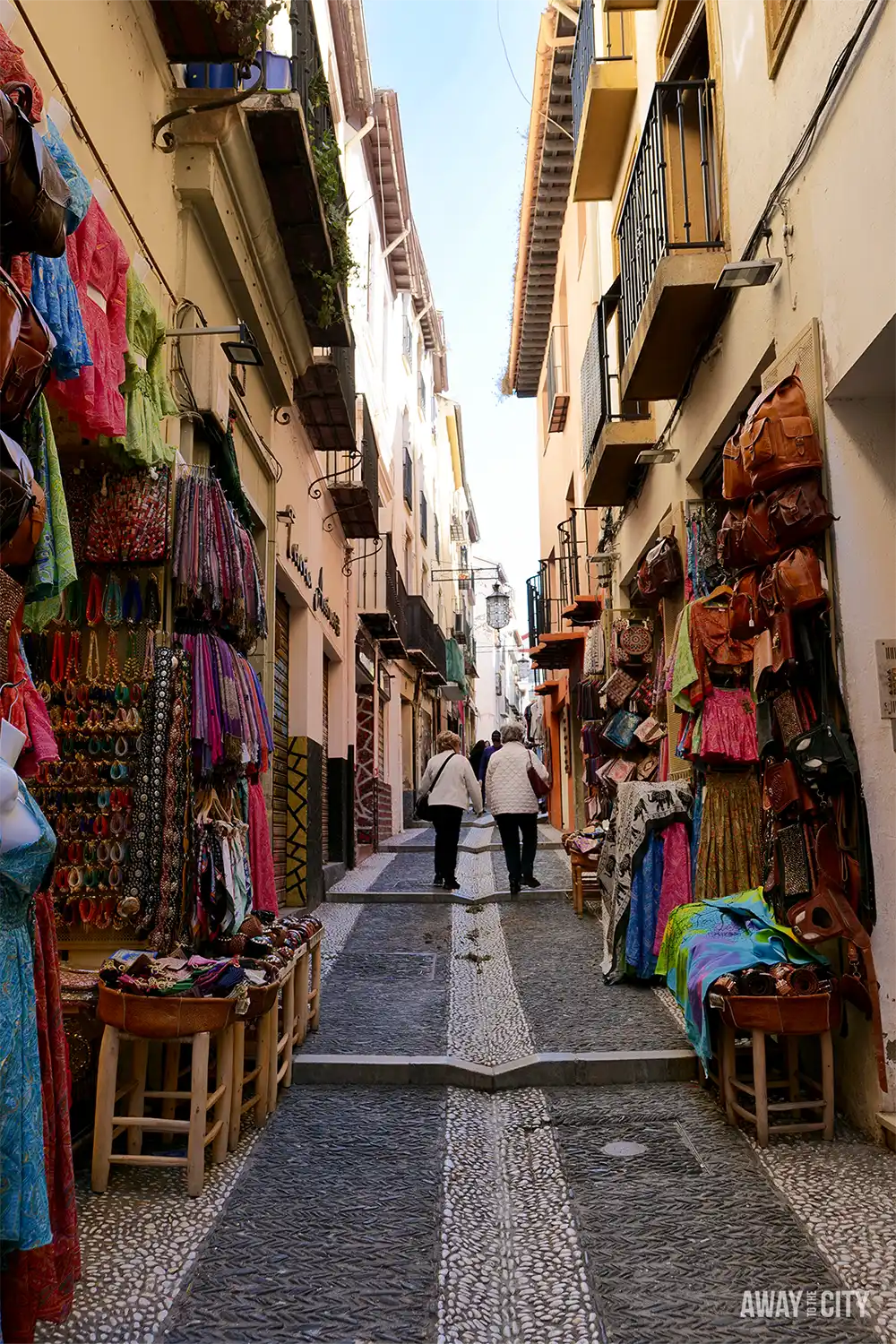 Narrow street in Granada’s Calle Caldereria Nueva, Little Morocco, lined with colourful textiles, hanging lanterns, and small shopfronts beneath tall buildings.