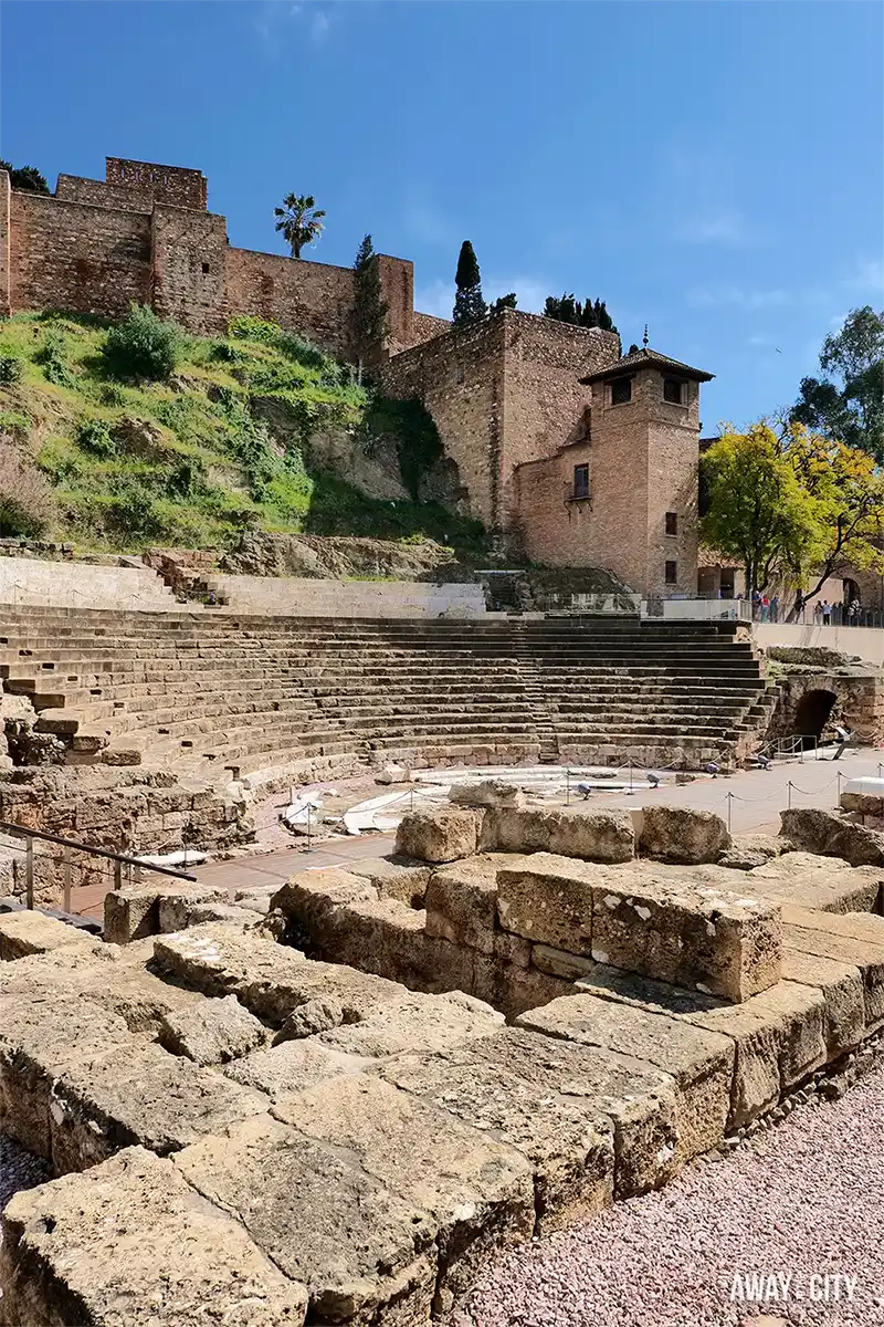 Ancient Roman theatre ruins with stepped stone seating beneath a hillside fortress, surrounded by weathered walls, greenery, and clear blue sky.