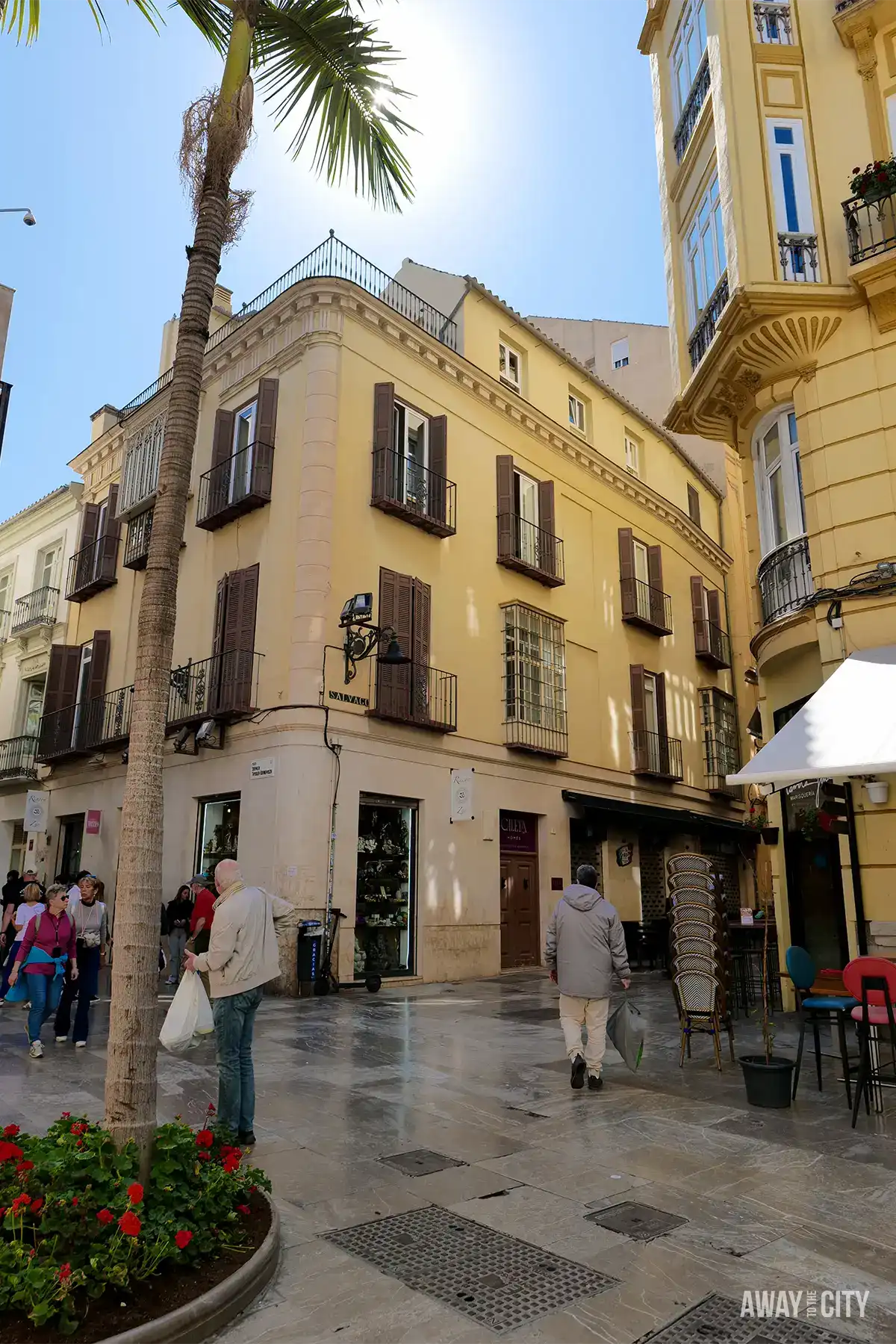 Corner of pastel yellow buildings with balconies and shutters in Malaga, palm tree overhead, and pedestrians walking across a bright cobbled square.