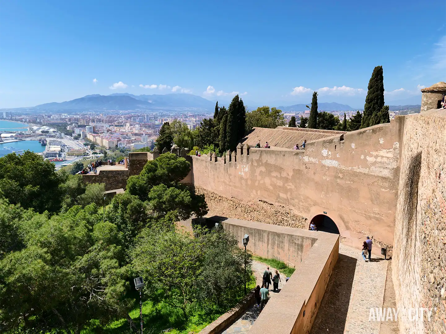 Panoramic view from Castillo de Gibralfaro overlooking Malaga city, harbour and mountains, with cypress trees and visitors along sunlit ramparts.
