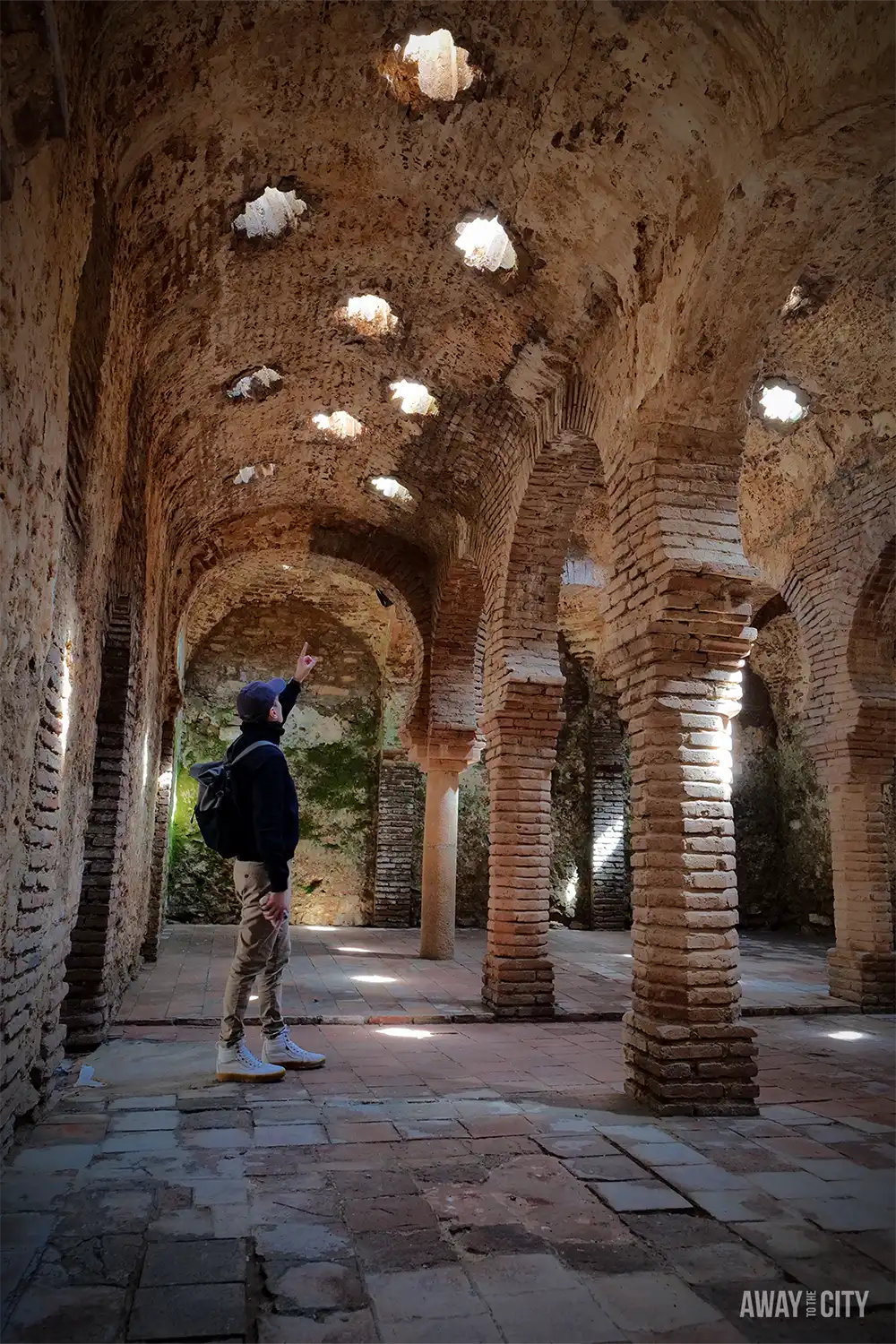 Interior of the Arab Baths in Ronda, featuring brick arches, stone columns, and a vaulted ceiling dotted with star-shaped skylights.