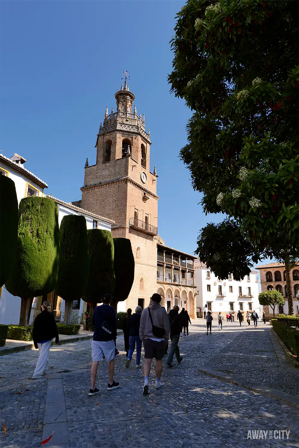 Plaza Duquesa de Parcent in Ronda old town, with historic church tower, trimmed trees, and people walking across a sunlit square.