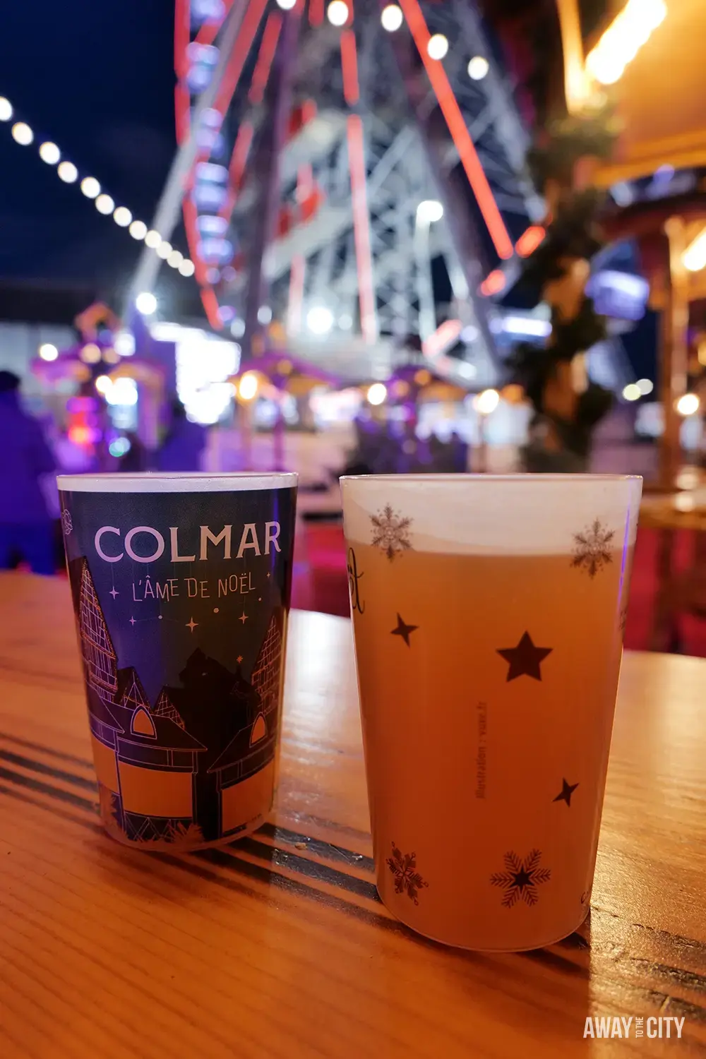 Close-up of festive drinks in cups on a wooden counter with blurred colourful lights and market atmosphere in the background.