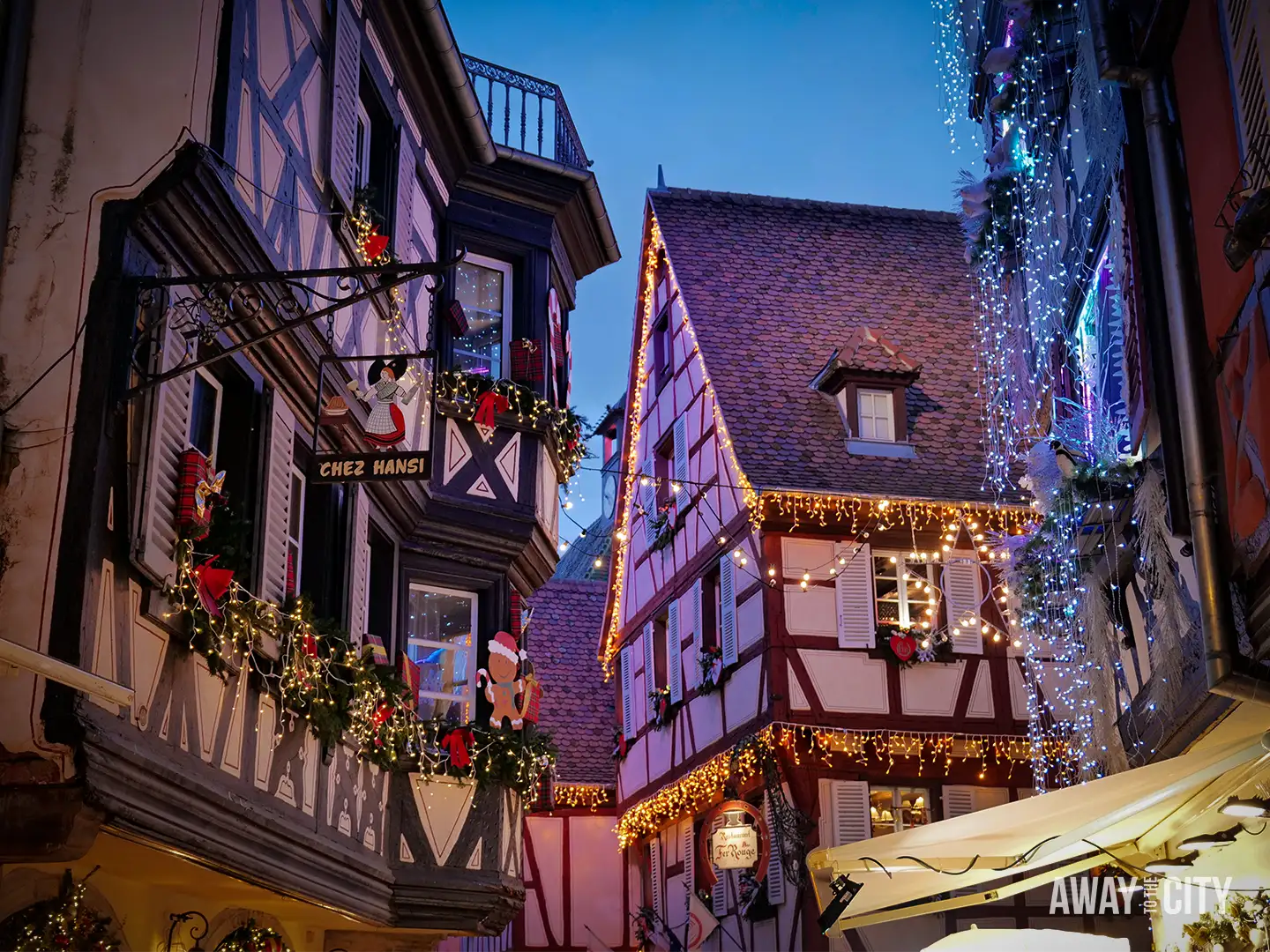 Charming Colmar street lined with traditional half-timbered houses, decorated with lights and seasonal ornaments in a cosy evening setting.