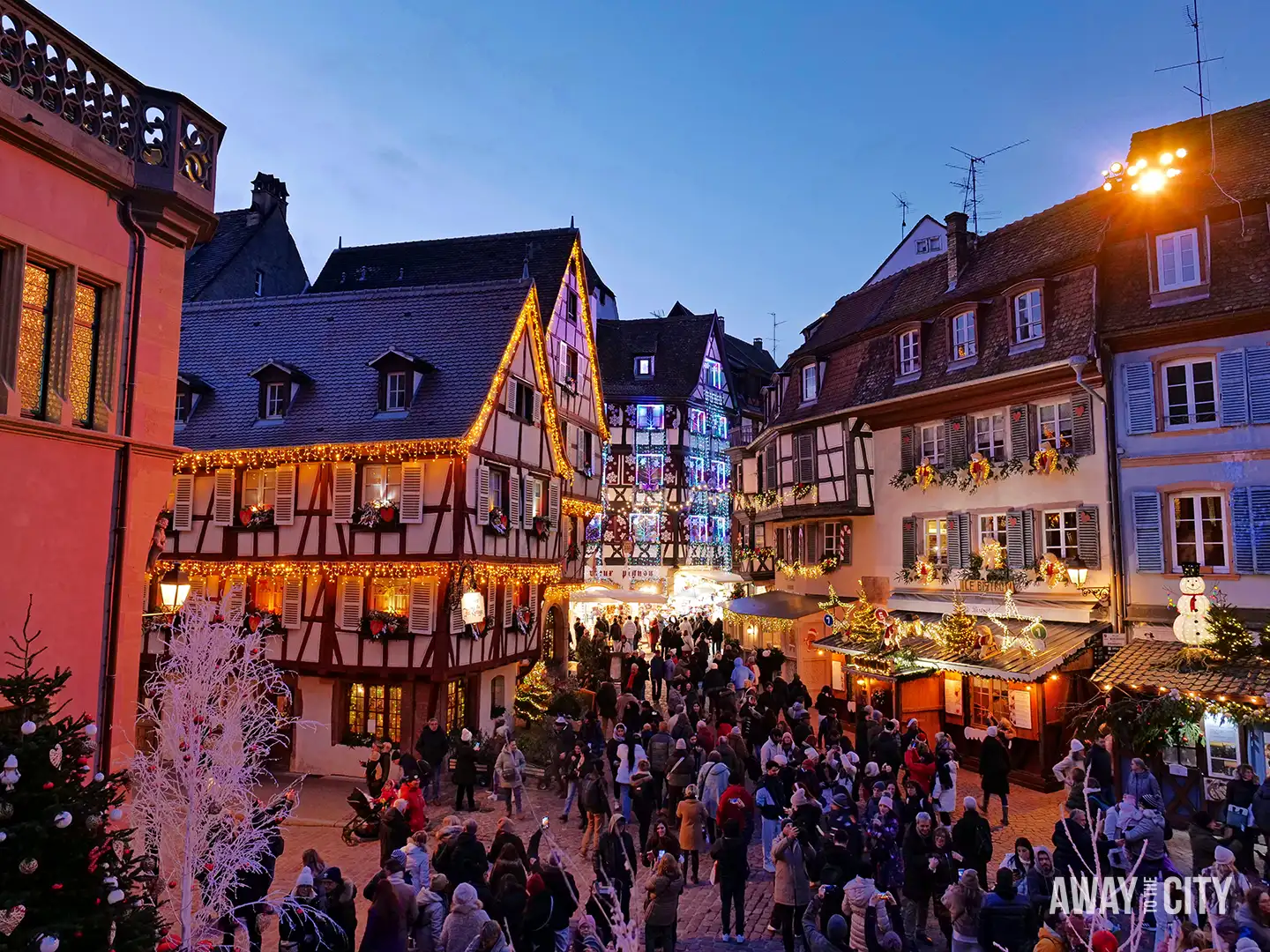 Busy Christmas market square in Colmar with half-timbered houses, festive lights, and crowds of people filling the street at dusk.
