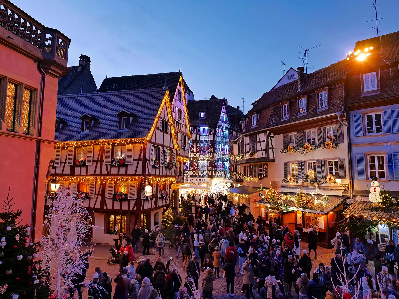 Busy Christmas market square in Colmar with half-timbered houses, festive lights, and crowds of people filling the street at dusk.
