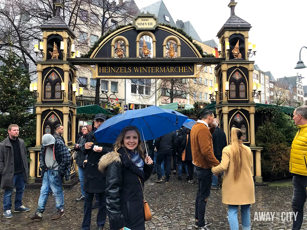 Outdoor market entrance in Cologne with decorative archway and people walking across a cobbled square on a cloudy day.