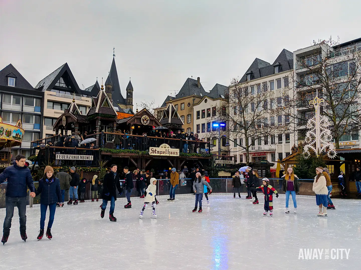 Open square in Cologne with market stalls and surrounding buildings, people gathered on an ice rink under overcast skies.
