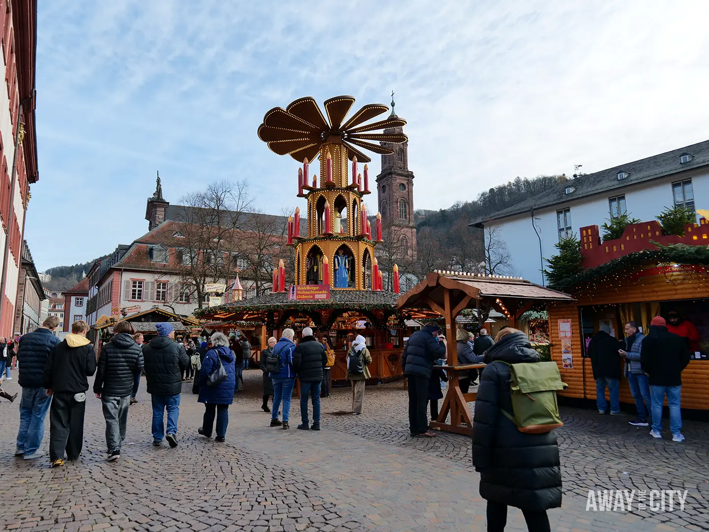 Christmas market square in Heidelberg with wooden stalls, a central pyramid decoration, and visitors walking across a cobbled open space.