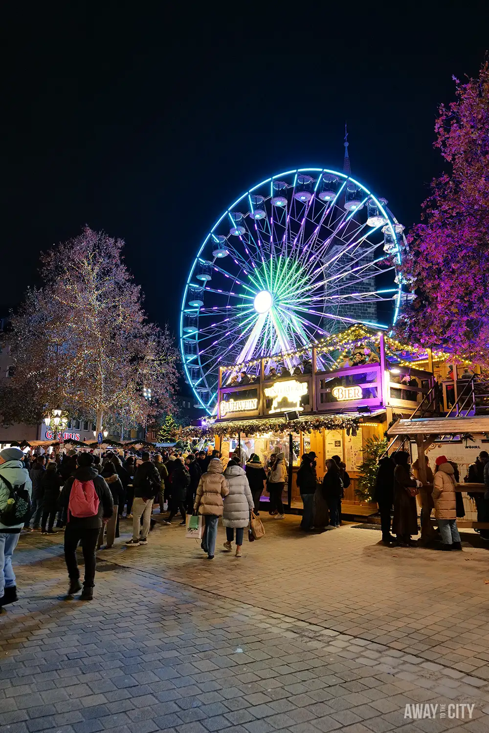 Nighttime Christmas market in Nuremberg with a brightly lit Ferris wheel, festive stalls, and crowds gathered under dark skies.