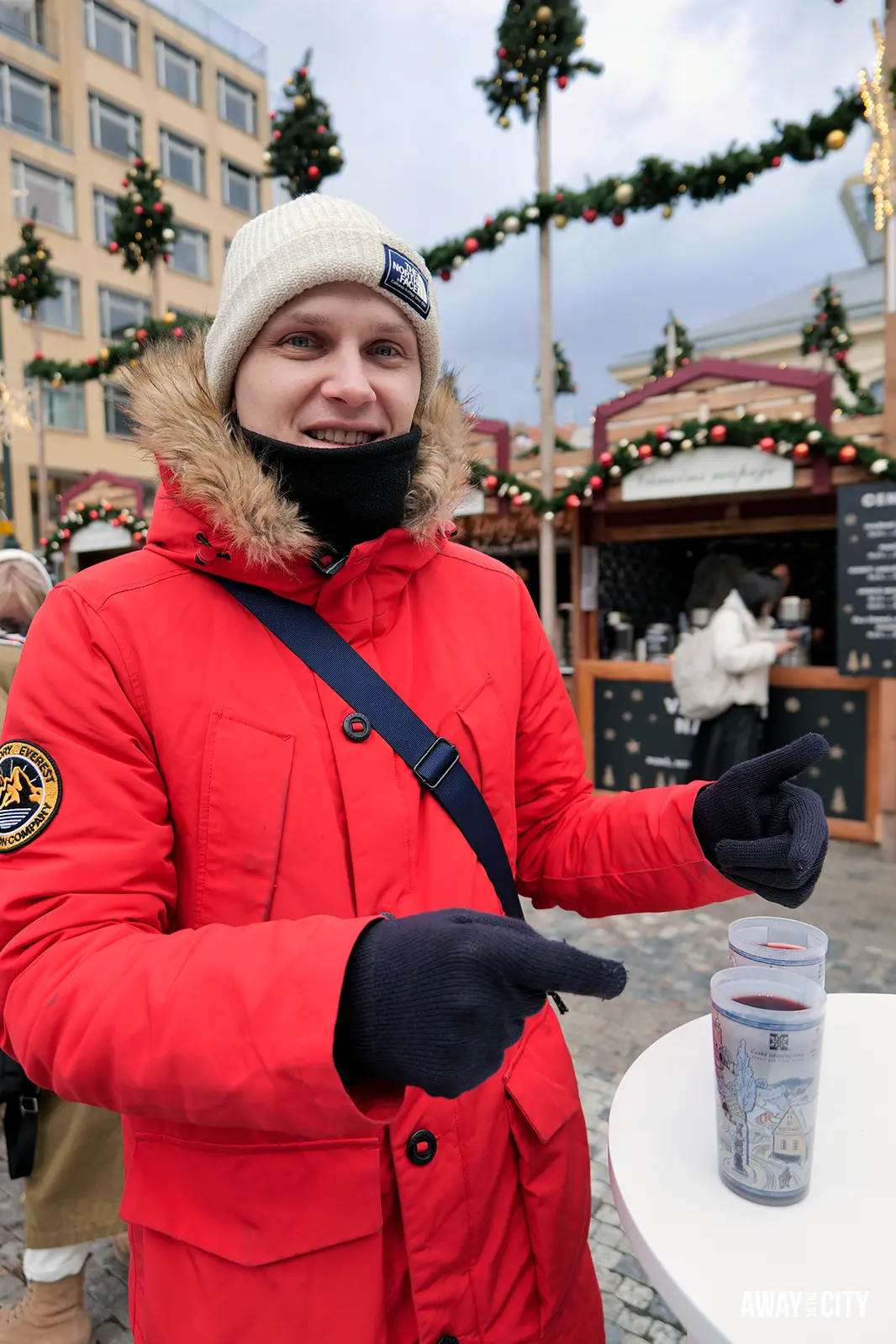 Person in a red coat holding a drink at a festive Prague market stall decorated with greenery and lights in winter.