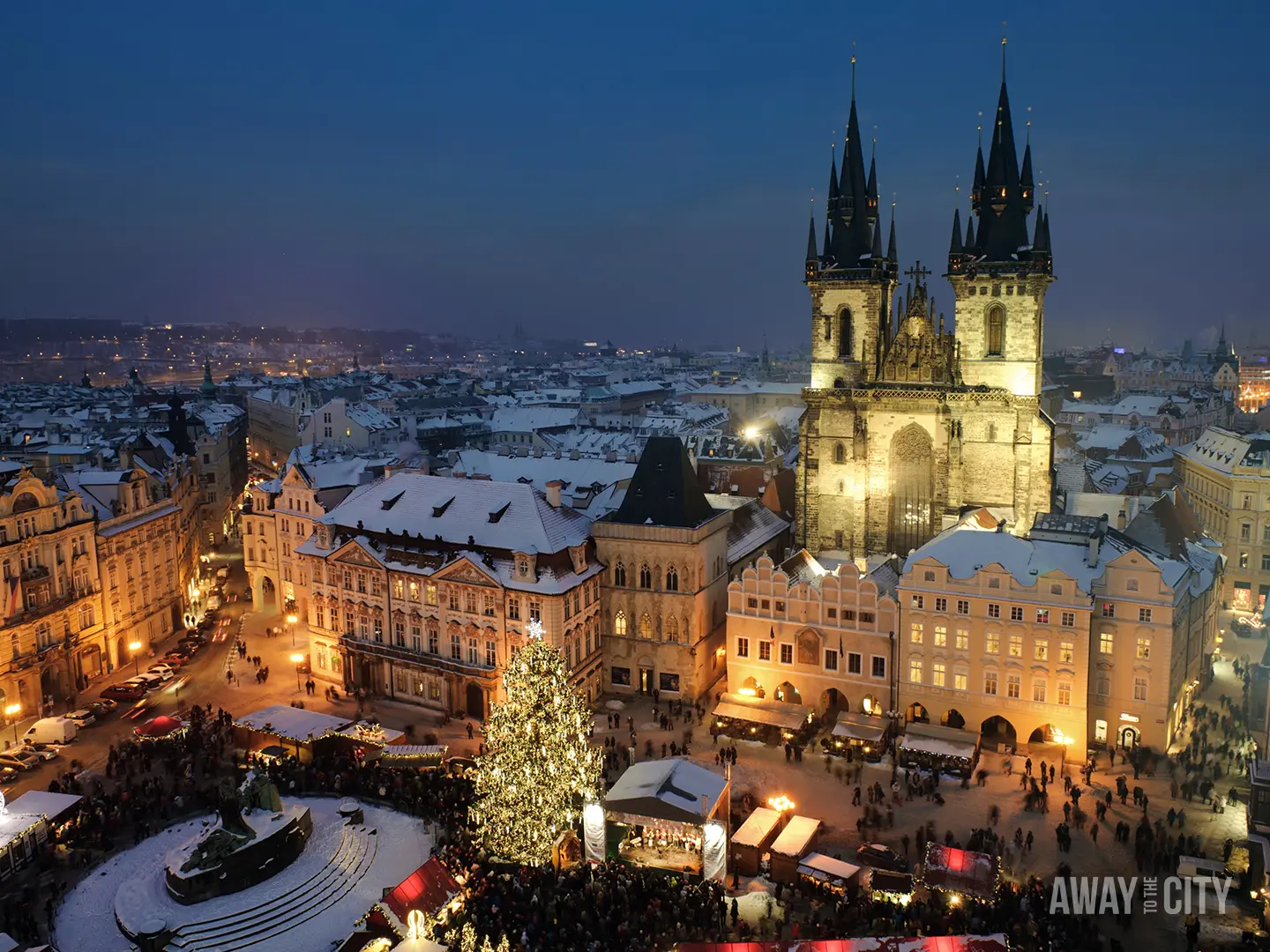 Elevated view of Prague’s Old Town Square at dusk with illuminated buildings, a large Christmas tree, and church spires above the city.