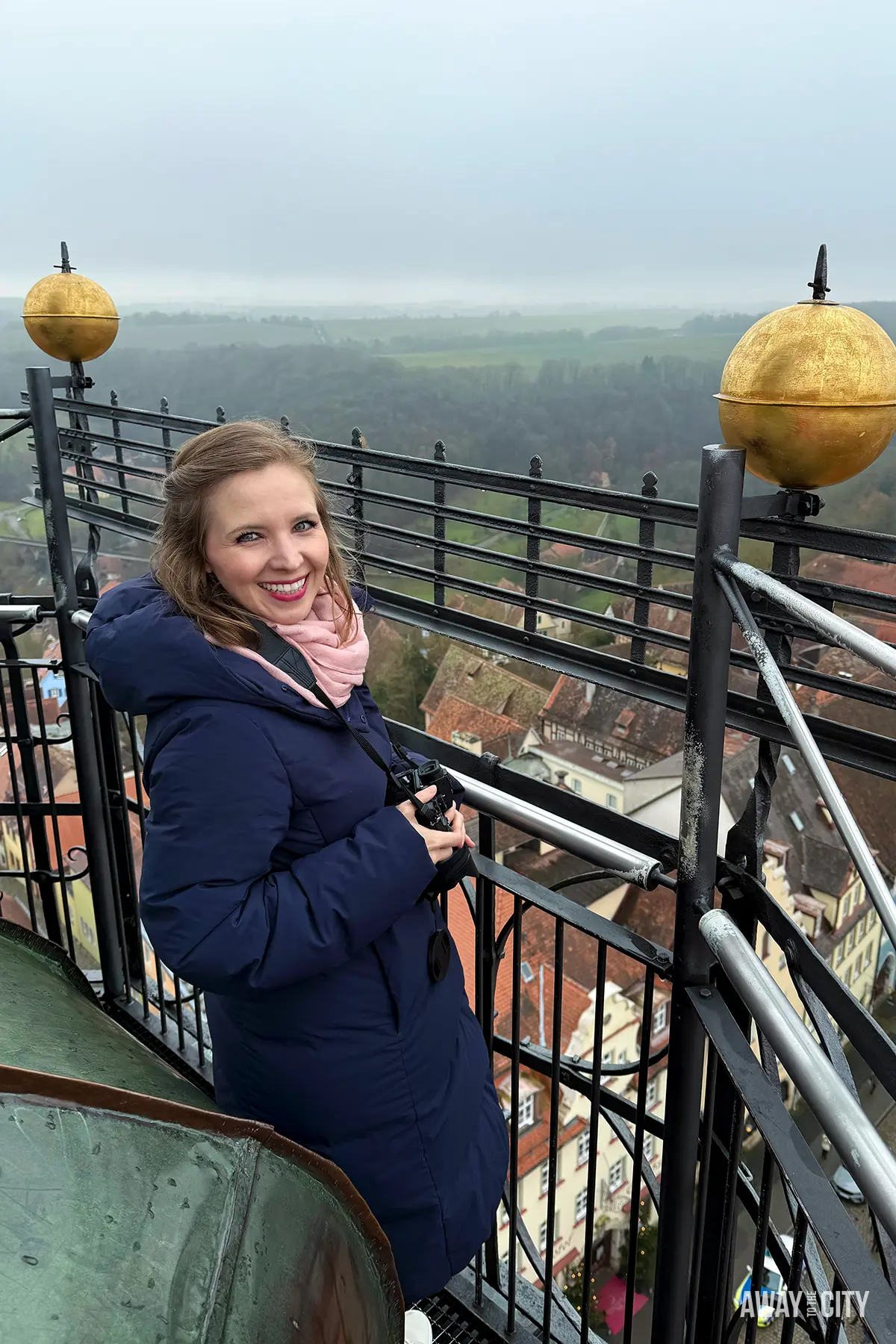 Woman standing on a tower balcony in Rothenburg overlooking a misty town landscape with rooftops and countryside stretching into the distance.