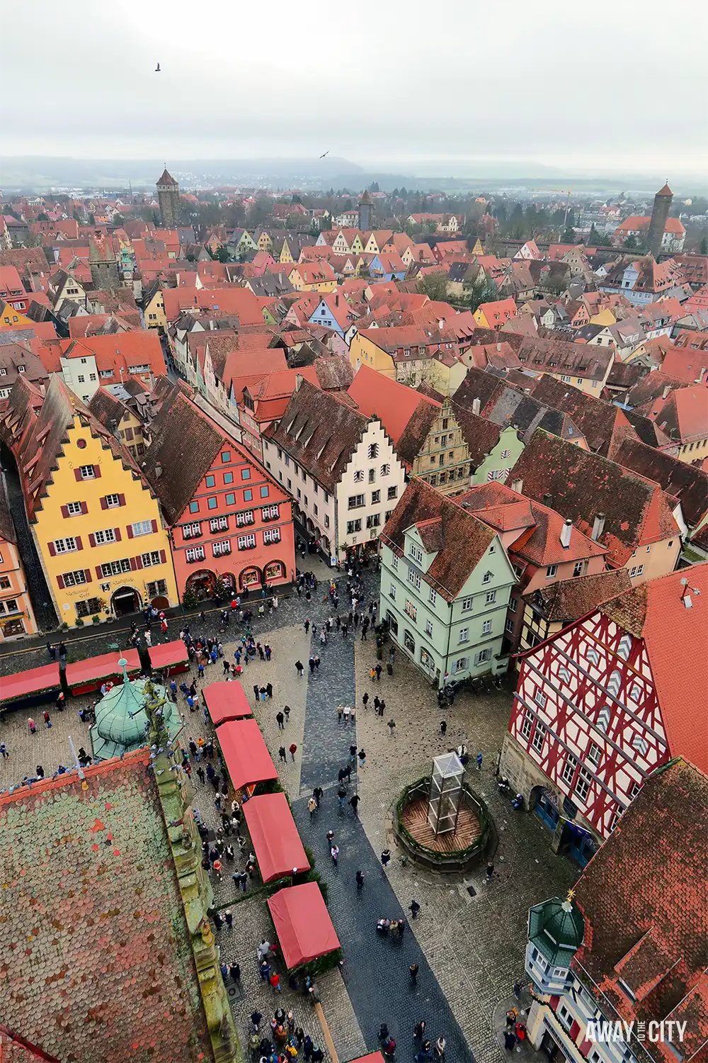 High view over Rothenburg showing dense red-roofed buildings, narrow streets, and a central square with people gathered below.