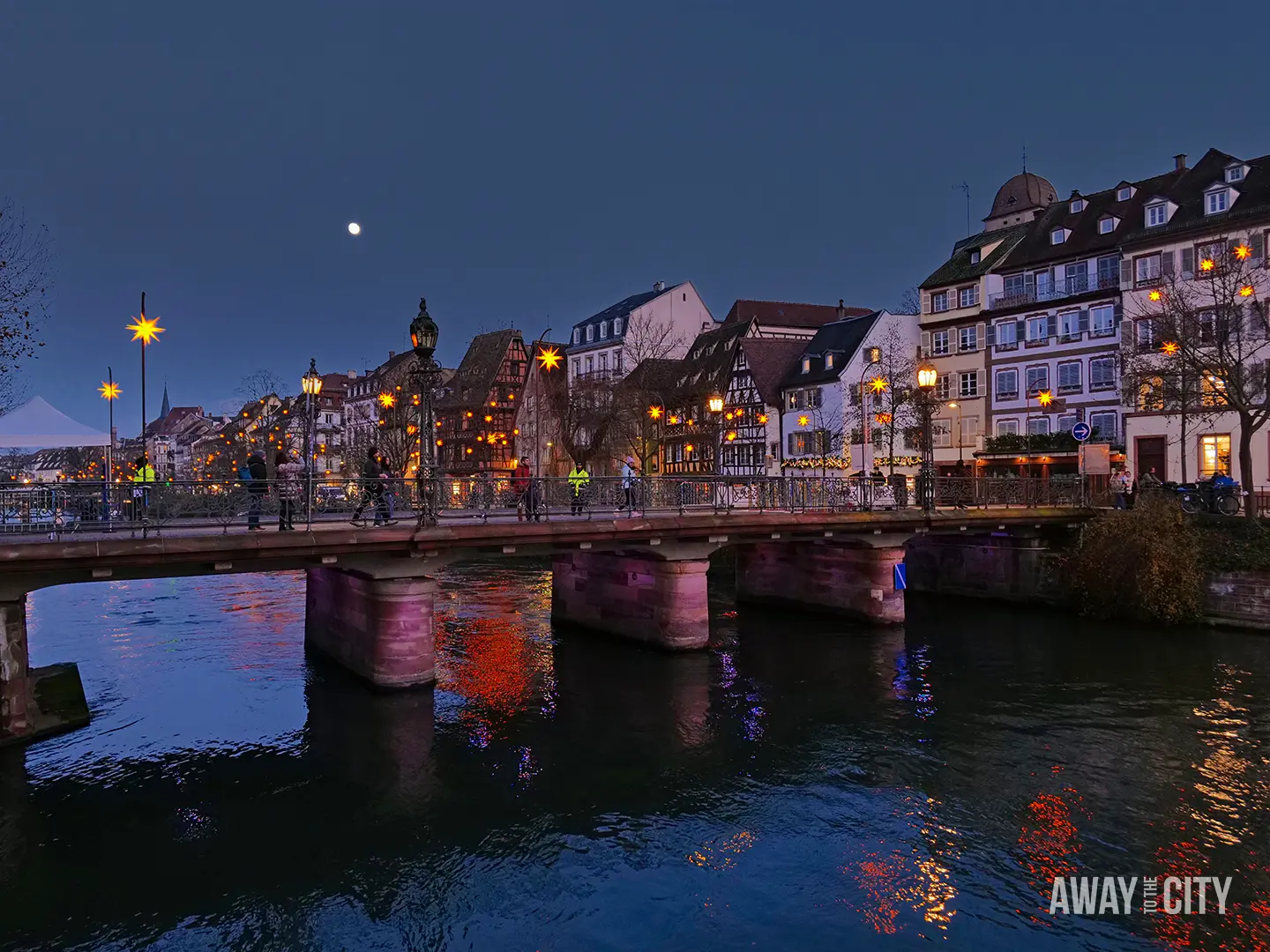 Evening view of Strasbourg with a lit bridge over the river, historic buildings along the water, and reflections shimmering on the surface.
