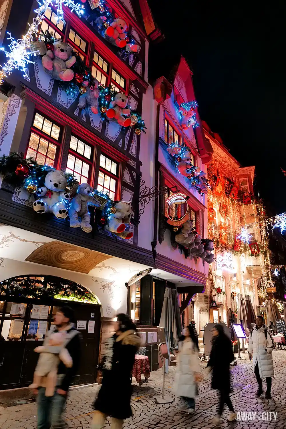 Festively decorated street in Strasbourg at night with illuminated half-timbered buildings, hanging ornaments, and people walking along cobbled pavement.