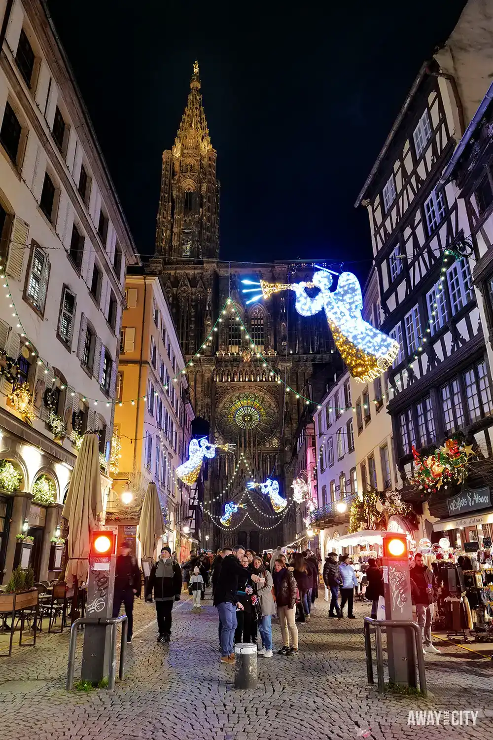 Night street in Strasbourg with illuminated decorations overhead, historic buildings lining the road, and people walking through a festive atmosphere.