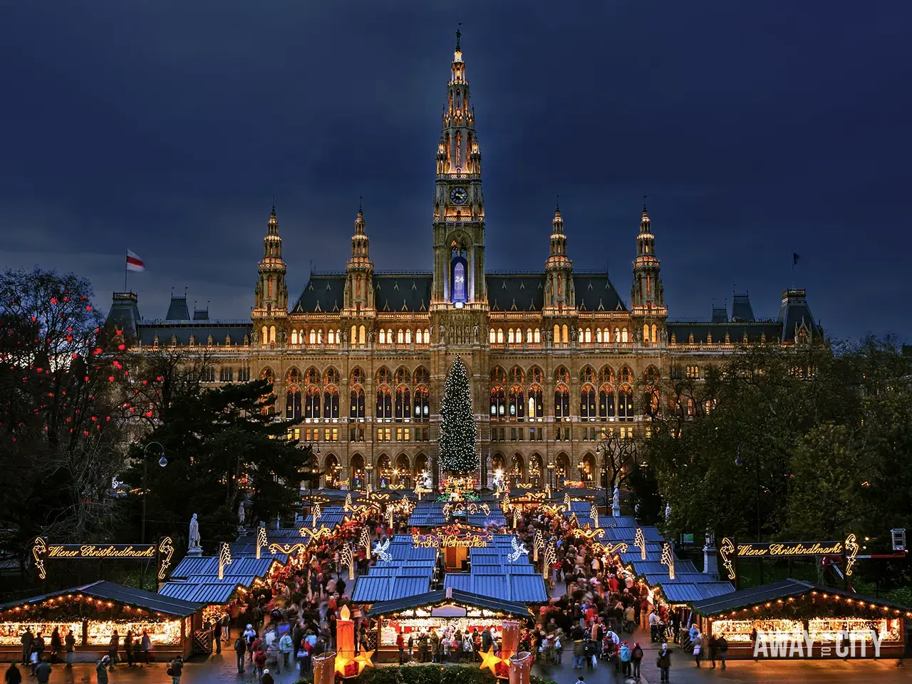 Grand illuminated building in Vienna with a large Christmas market in front, filled with stalls, lights, and crowds under a twilight sky.