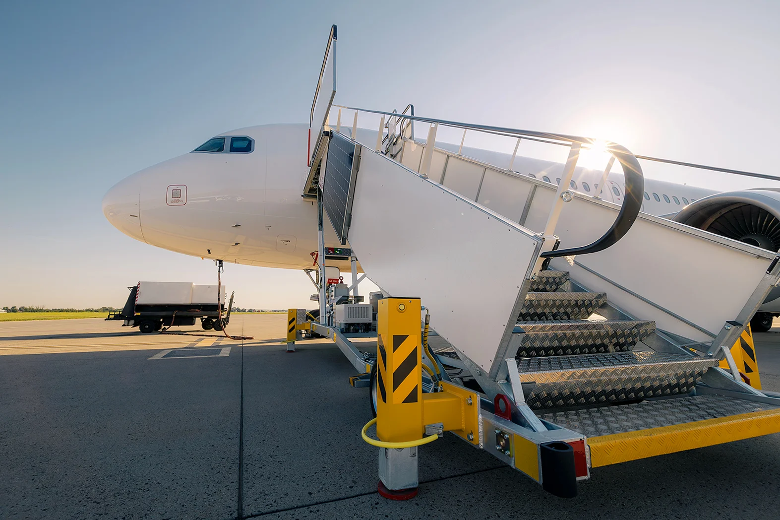 A white passenger plane is parked on the tarmac with a mobile boarding staircase connected and the sun shining.
