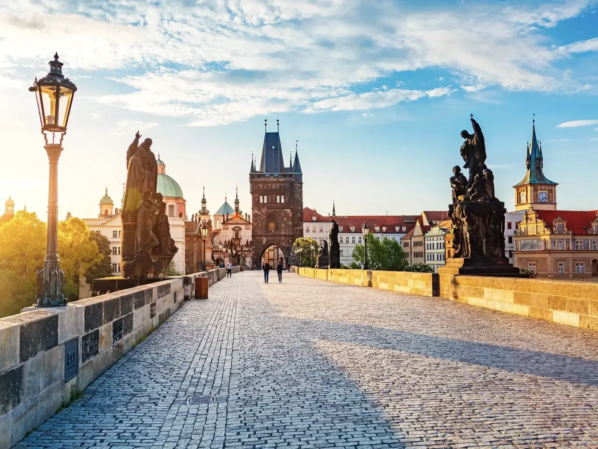 Prague's Charles Bridge, adorned with statues and streetlights, is bathed in sunlight, with the Old Town Bridge Tower and city skyline in the background.