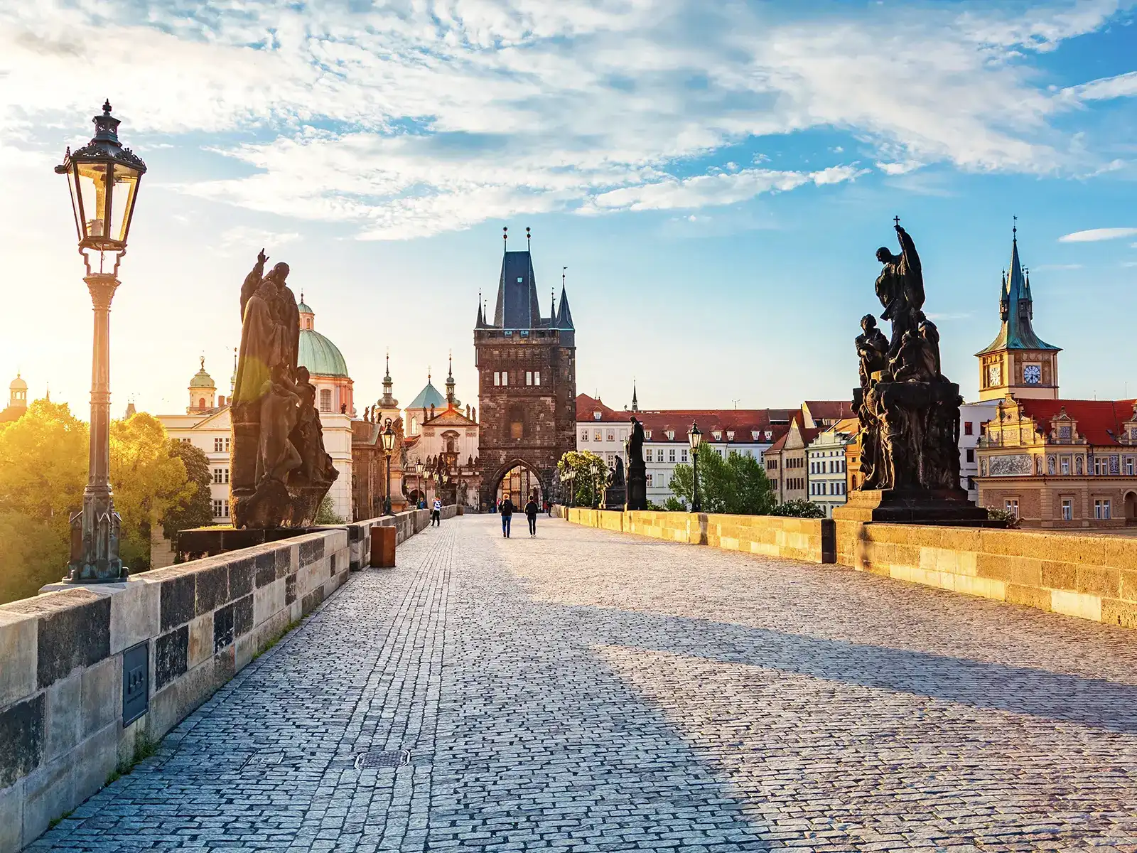 Prague's Charles Bridge, adorned with statues and streetlights, is bathed in sunlight, with the Old Town Bridge Tower and city skyline in the background.