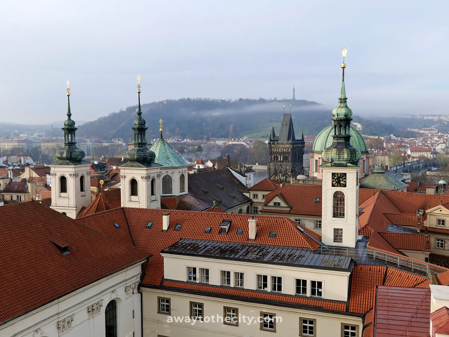 A stunning view of Prague's red rooftops and historic spires from the Klementinum, with a misty hill in the distance.