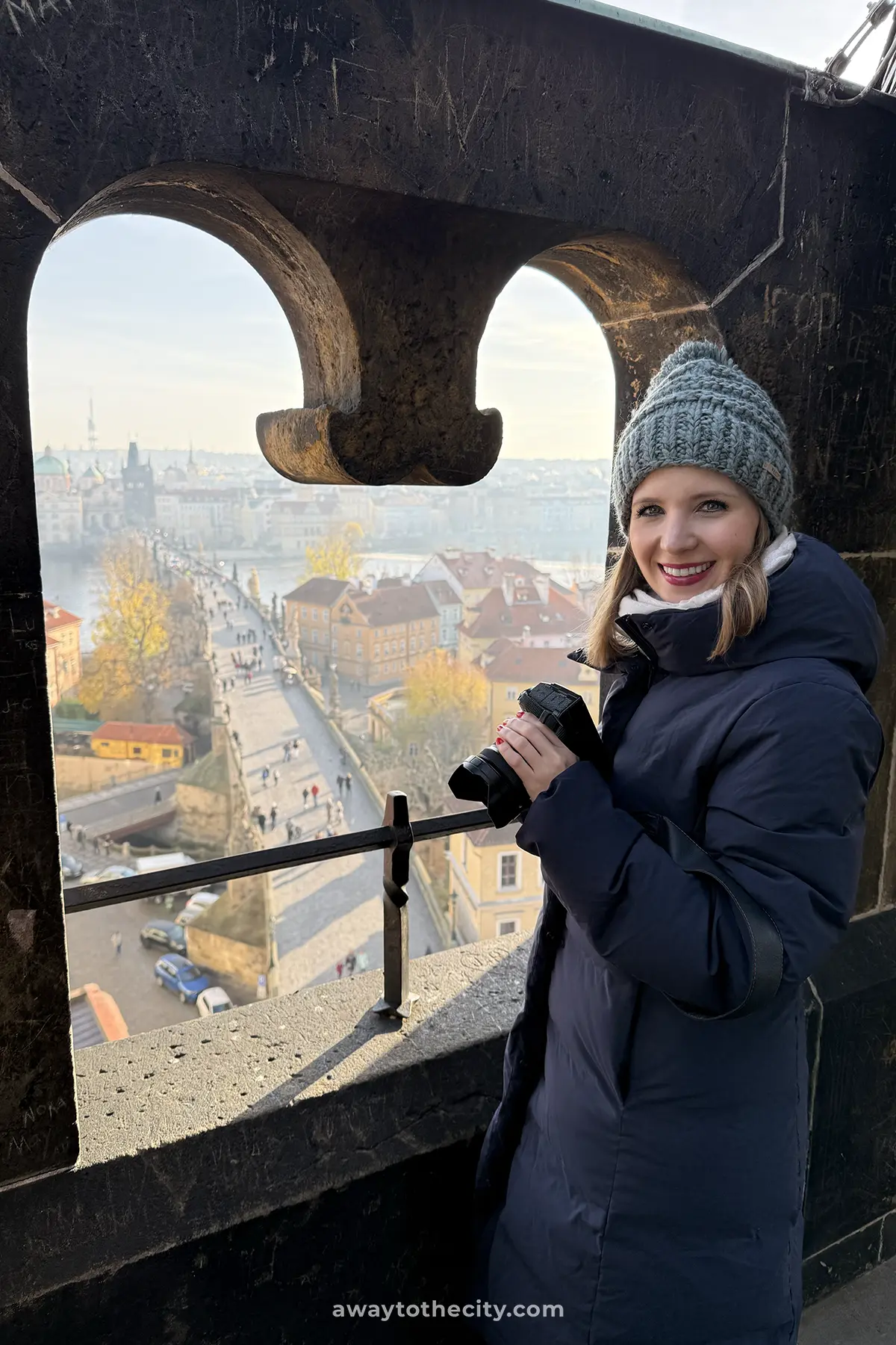 A smiling woman in a warm coat and hat holds a camera, overlooking Prague's Charles Bridge and cityscape from the Lesser Town Bridge Tower.
