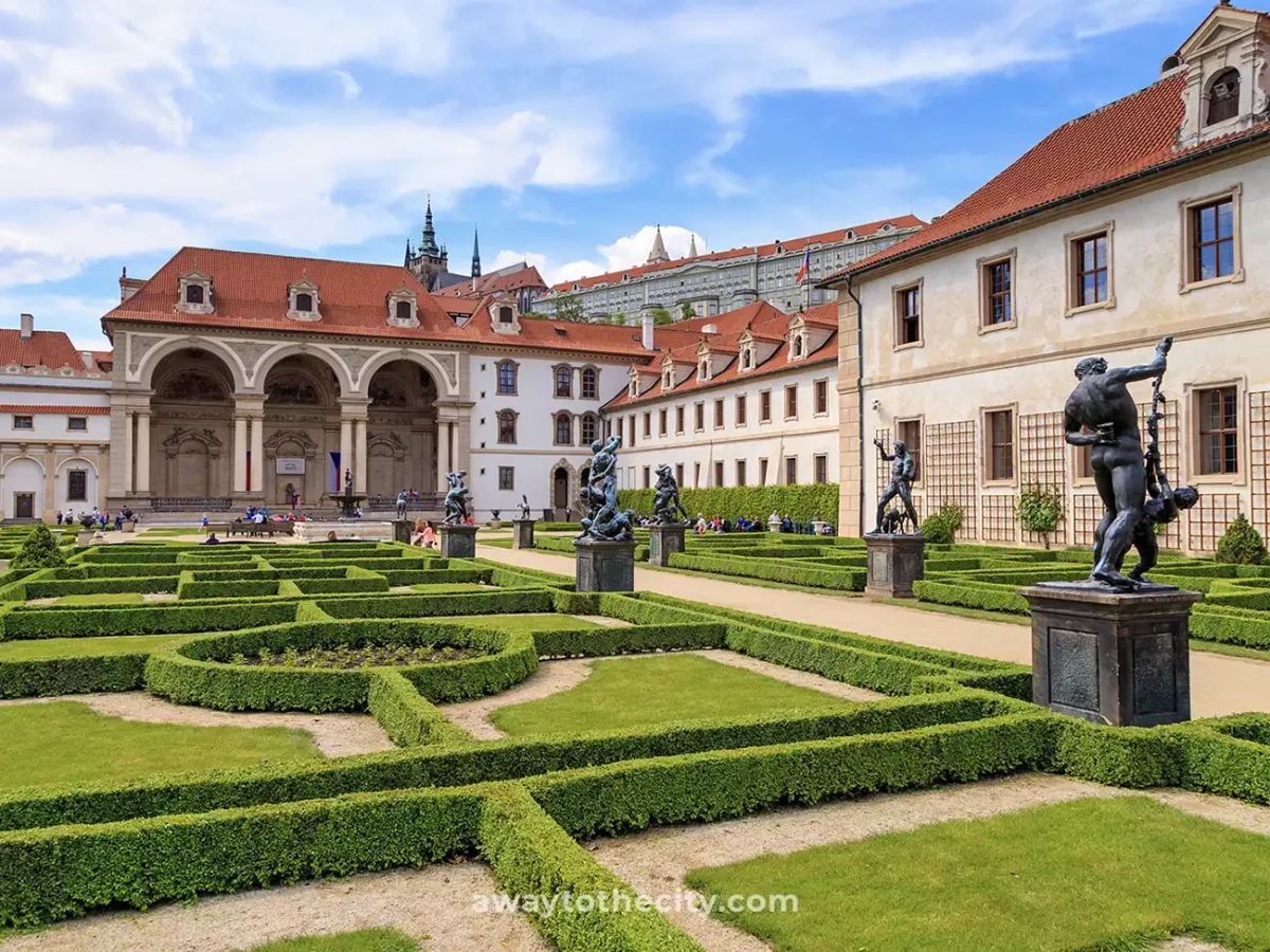 The formal Wallenstein Palace Gardens in Prague feature manicured hedges, classical statues, and grand palace buildings under a blue sky.
