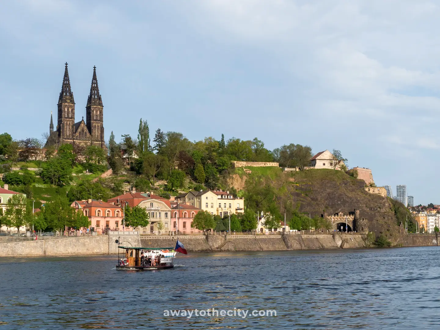 View across the Vltava River towards Vyšehrad’s hilltop fortress, with twin church spires, riverside buildings and a boat.