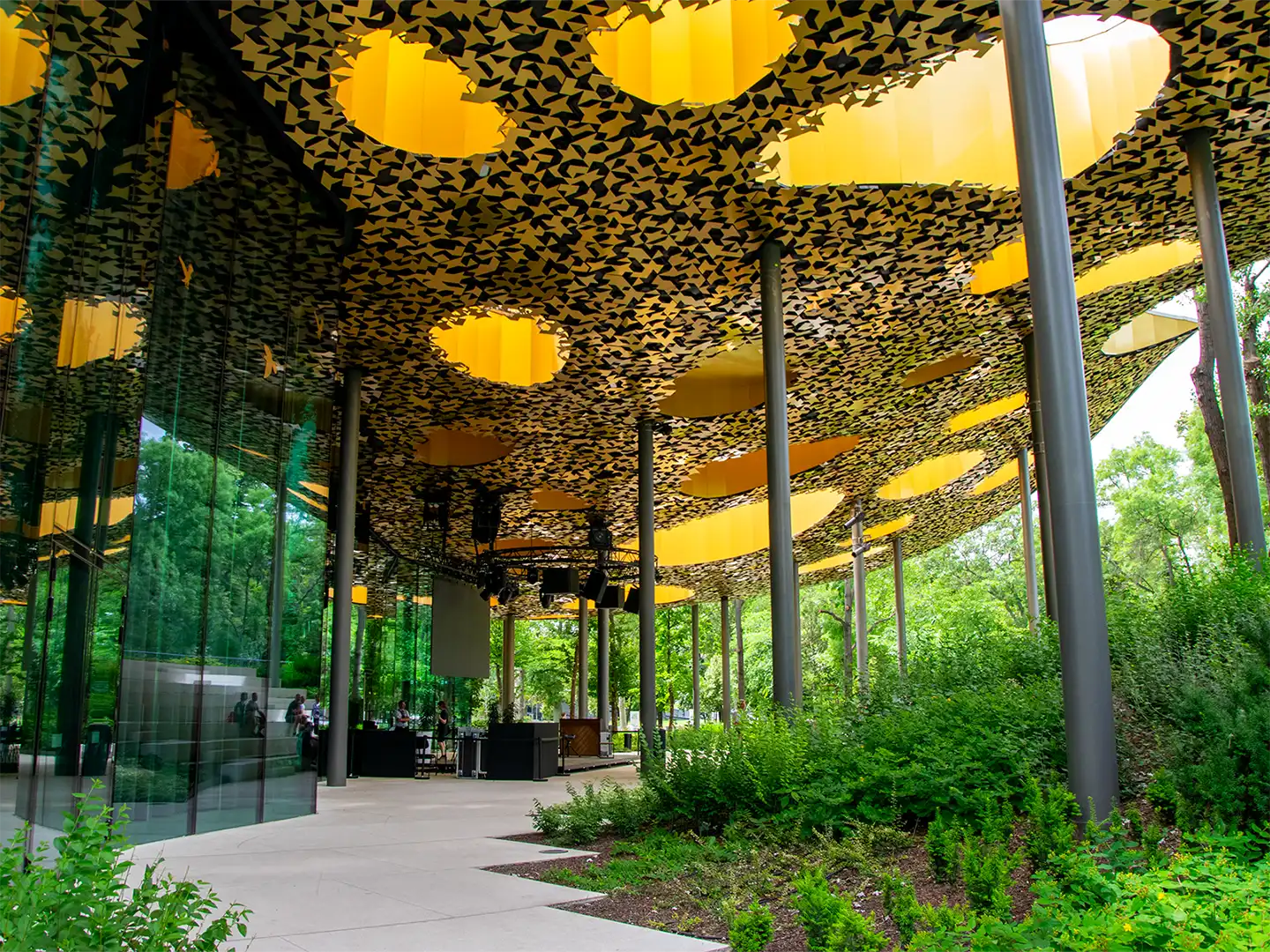 Exterior of the House of Music Hungary in Budapest, featuring a distinctive perforated golden roof, glass walls, slender columns, and surrounding parkland.